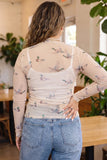 A woman viewed from behind wearing a beige sheer long-sleeve top with a multicolor duck print, layered over a white camisole, paired with blue jeans in a cafe setting.