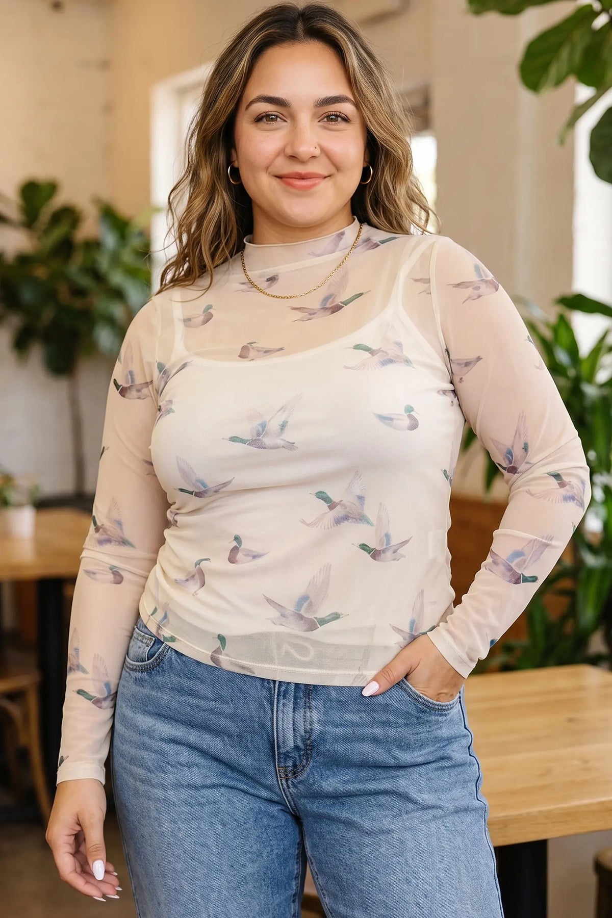 A woman wearing a sheer long-sleeve mock neck top with a light beige background and a delicate bird print, paired with blue jeans, standing in a softly lit indoor setting with plants and wooden furniture.