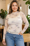 A woman wearing a sheer long-sleeve mock neck top with a light beige background and a delicate bird print, paired with blue jeans, standing in a softly lit indoor setting with plants and wooden furniture.
