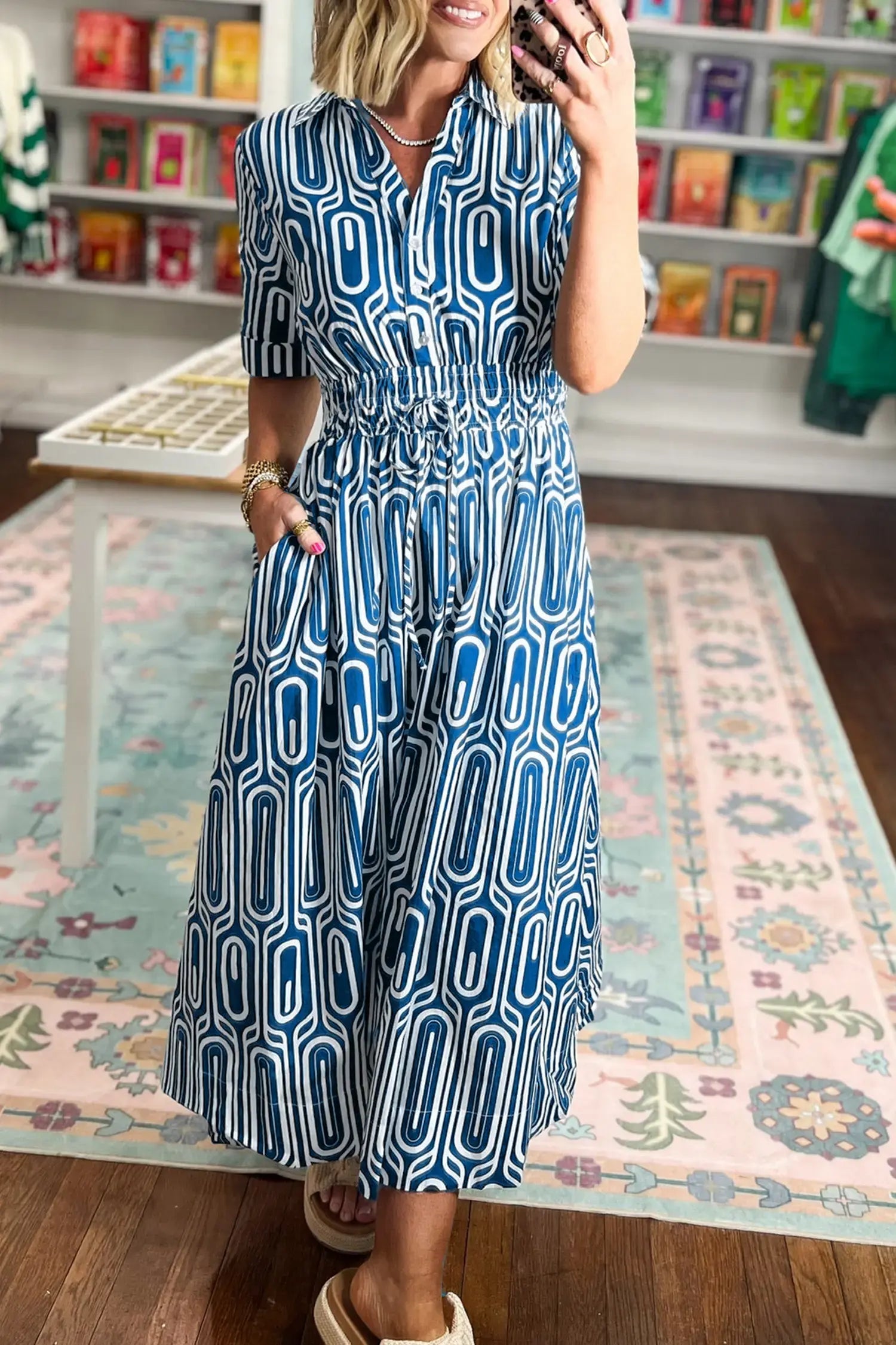 A woman wearing a blue and white geometric pattern midi dress with short sleeves, a collared neckline, and a smocked waist, standing in a retail store on a patterned rug.