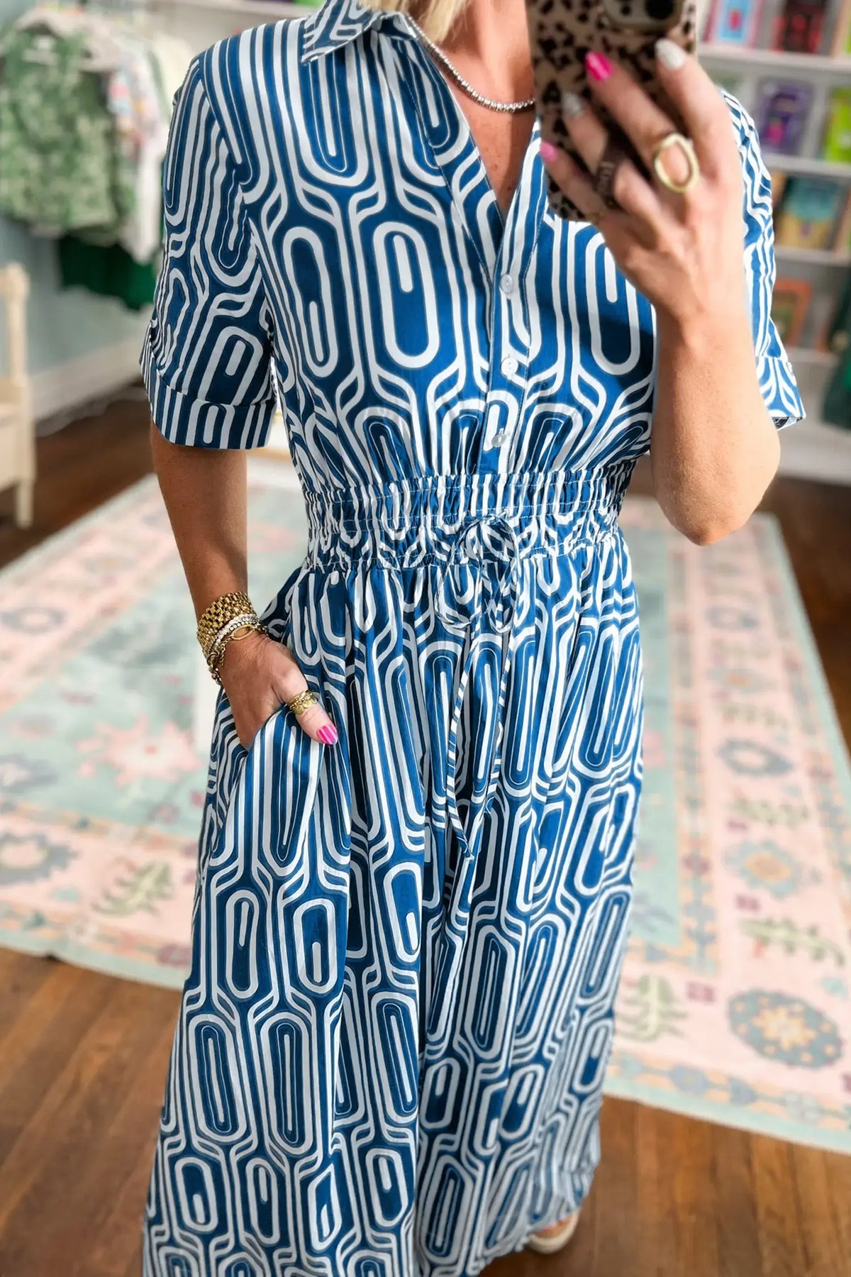 A woman takes a mirror selfie in a boutique wearing a blue and white geometric patterned midi dress with short sleeves, a collared neckline, and a smocked waist.