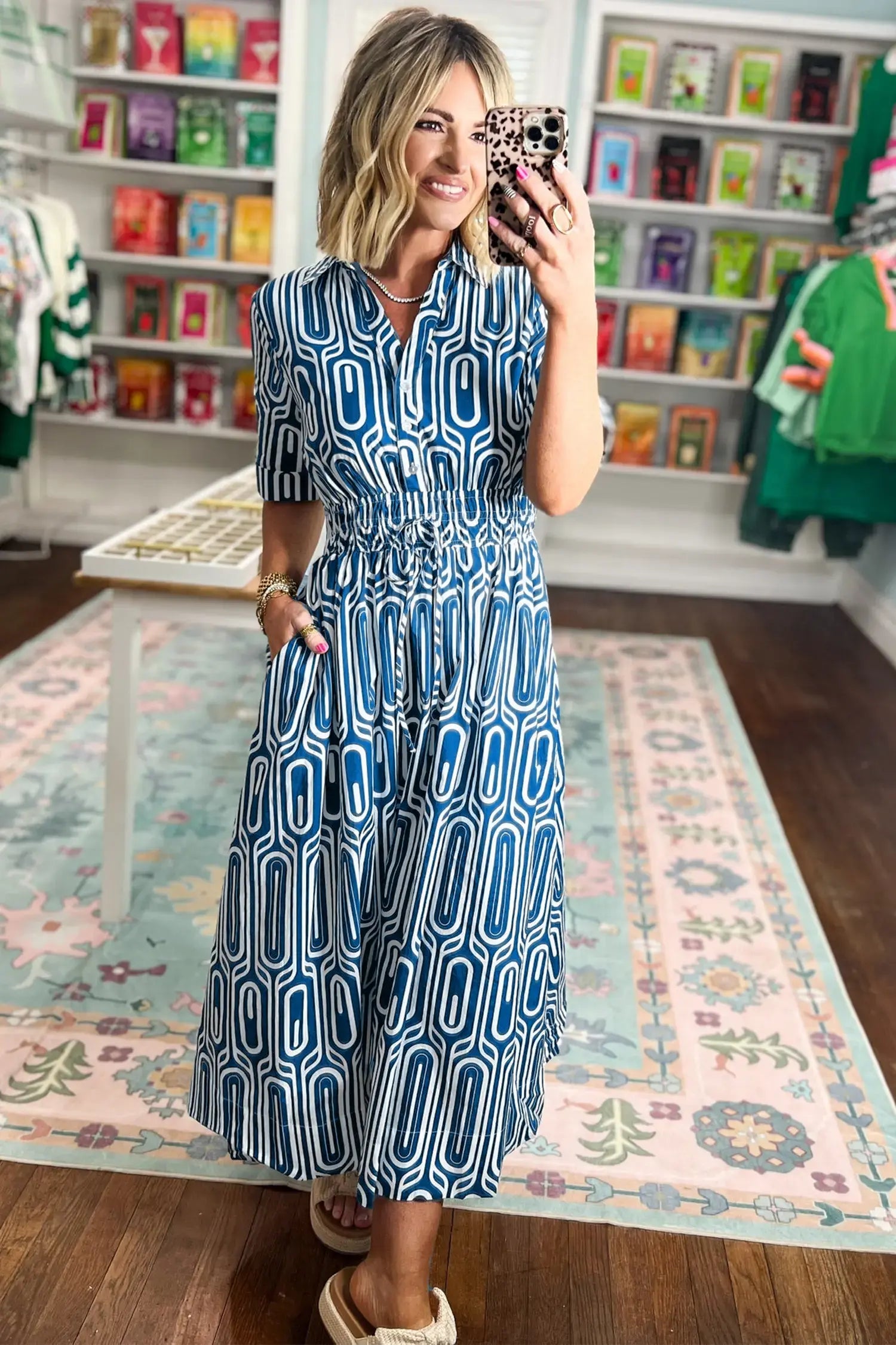 A woman takes a mirror selfie in a boutique while wearing a blue and white geometric pattern midi dress with short sleeves and a smocked waist.