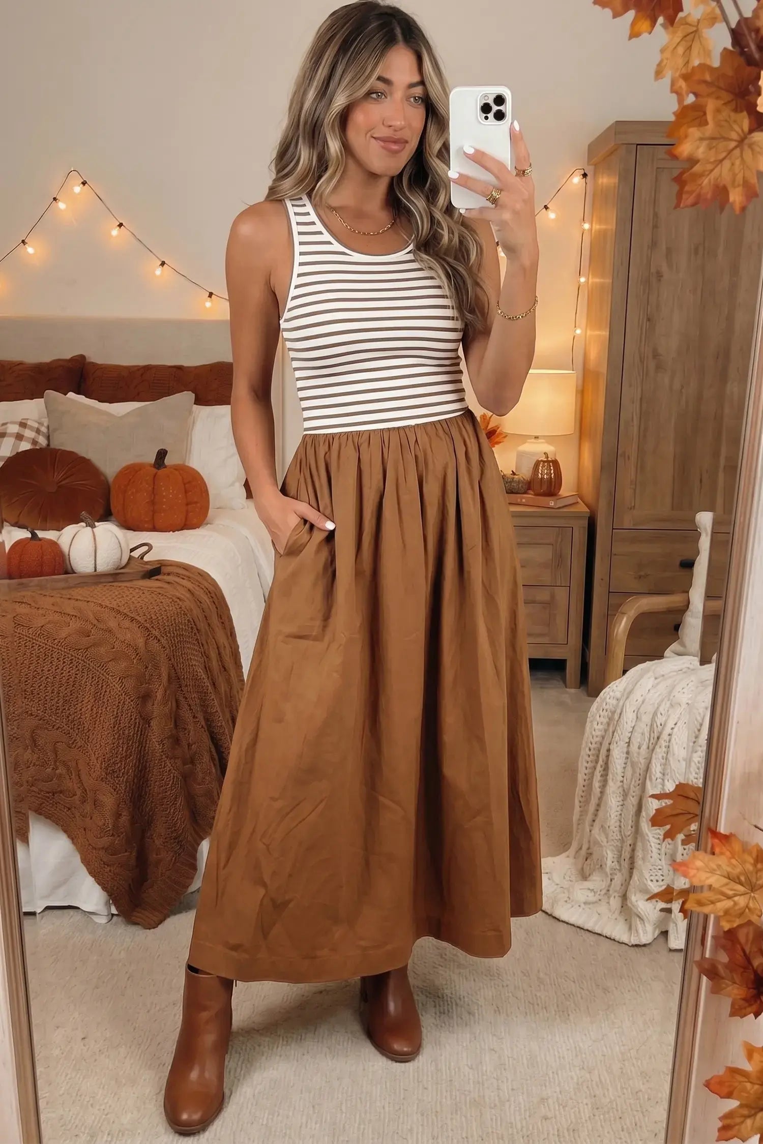 A woman takes a mirror selfie in a bedroom setting, wearing a sleeveless white tank top with thin brown horizontal stripes and a high-waisted, pleated brown maxi skirt with side pockets, paired with brown ankle boots.