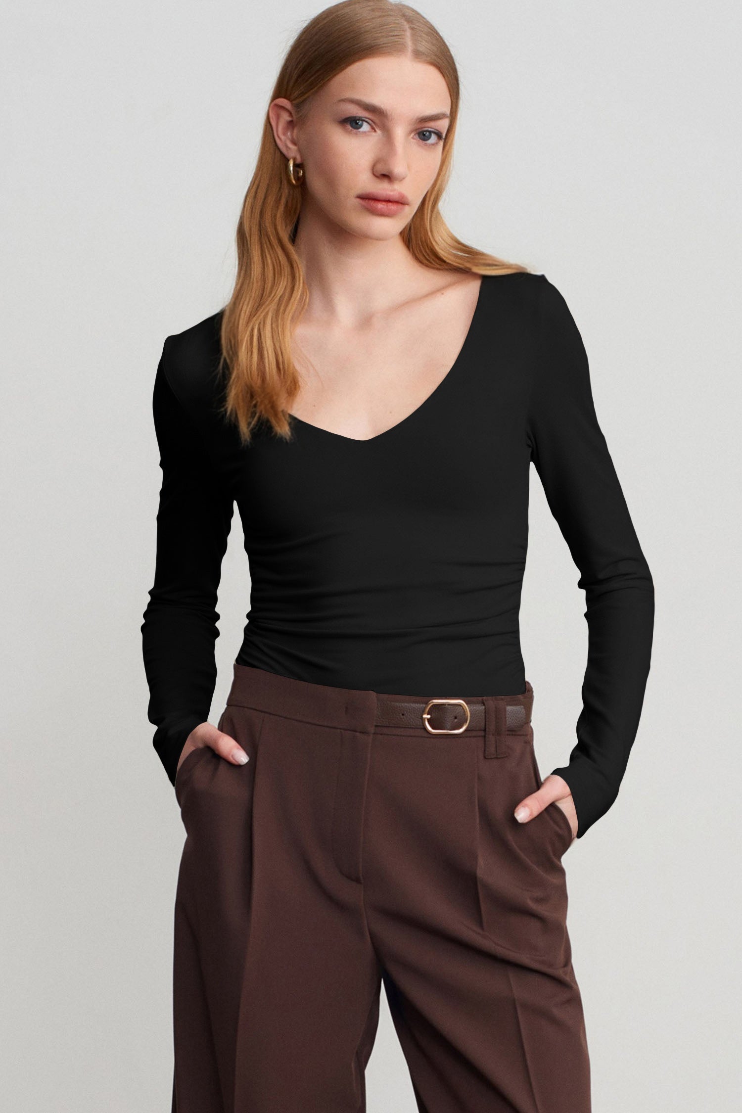 Woman in fitted black V-neck top with brown trousers against light gray background.