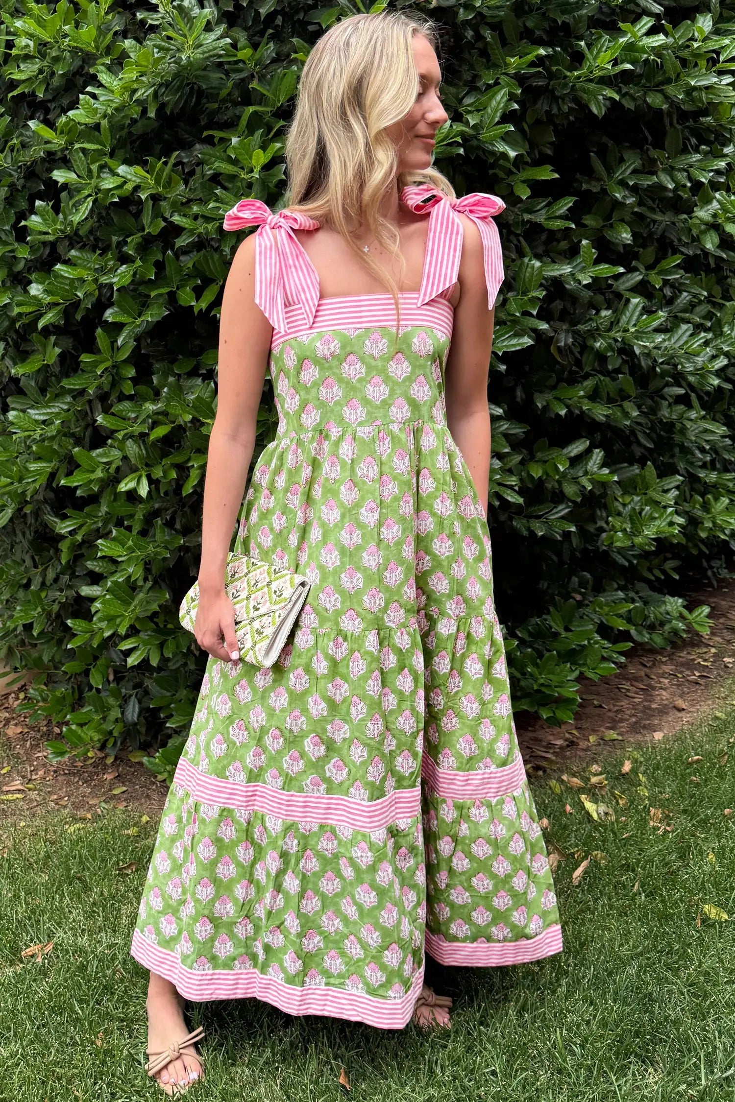 Woman wearing a green floral dress with pink trim standing in front of a bush.