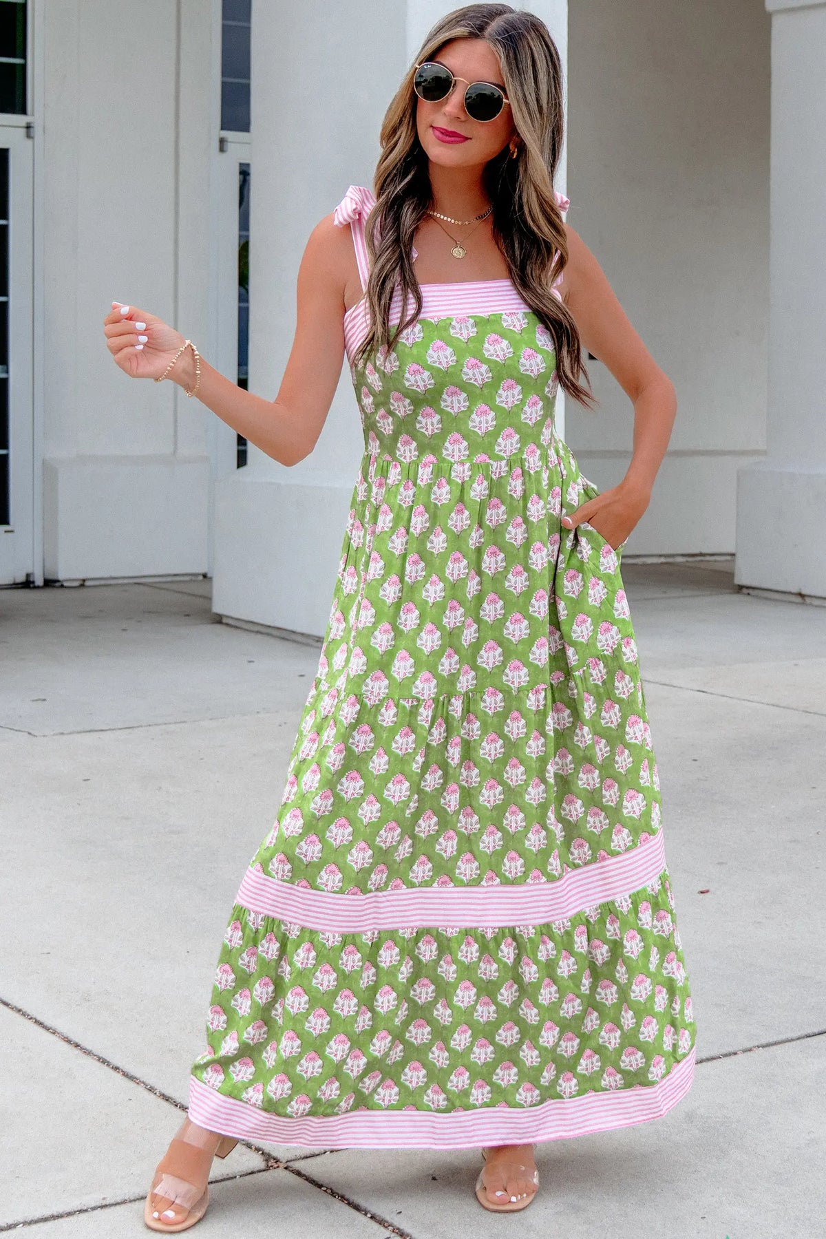 A woman modeling a green and pink floral print maxi dress with pink striped trim, standing outdoors in front of a white building.