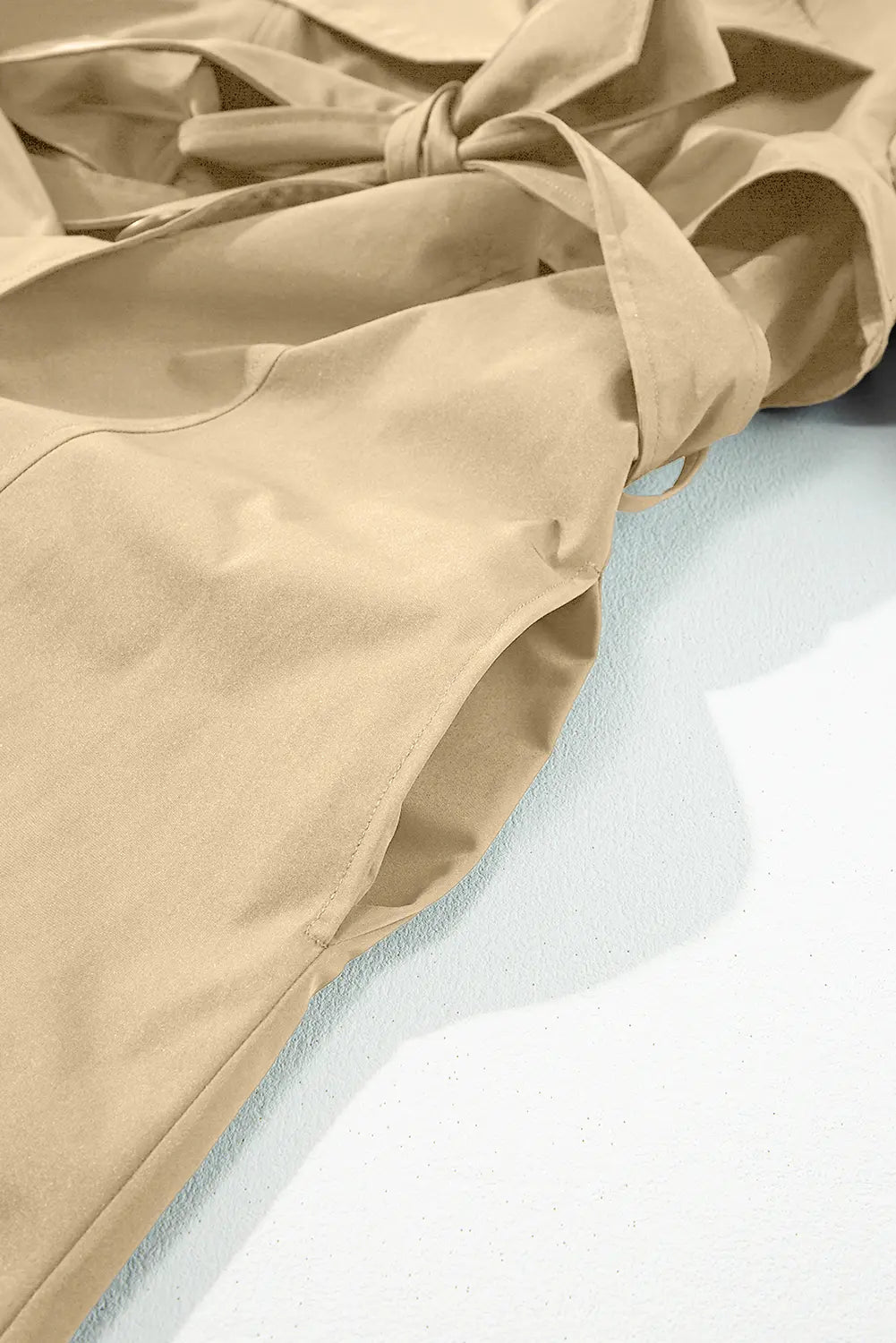 A close-up of a beige trench coat or wrap jacket with a tie belt, draped diagonally across a light blue surface.
