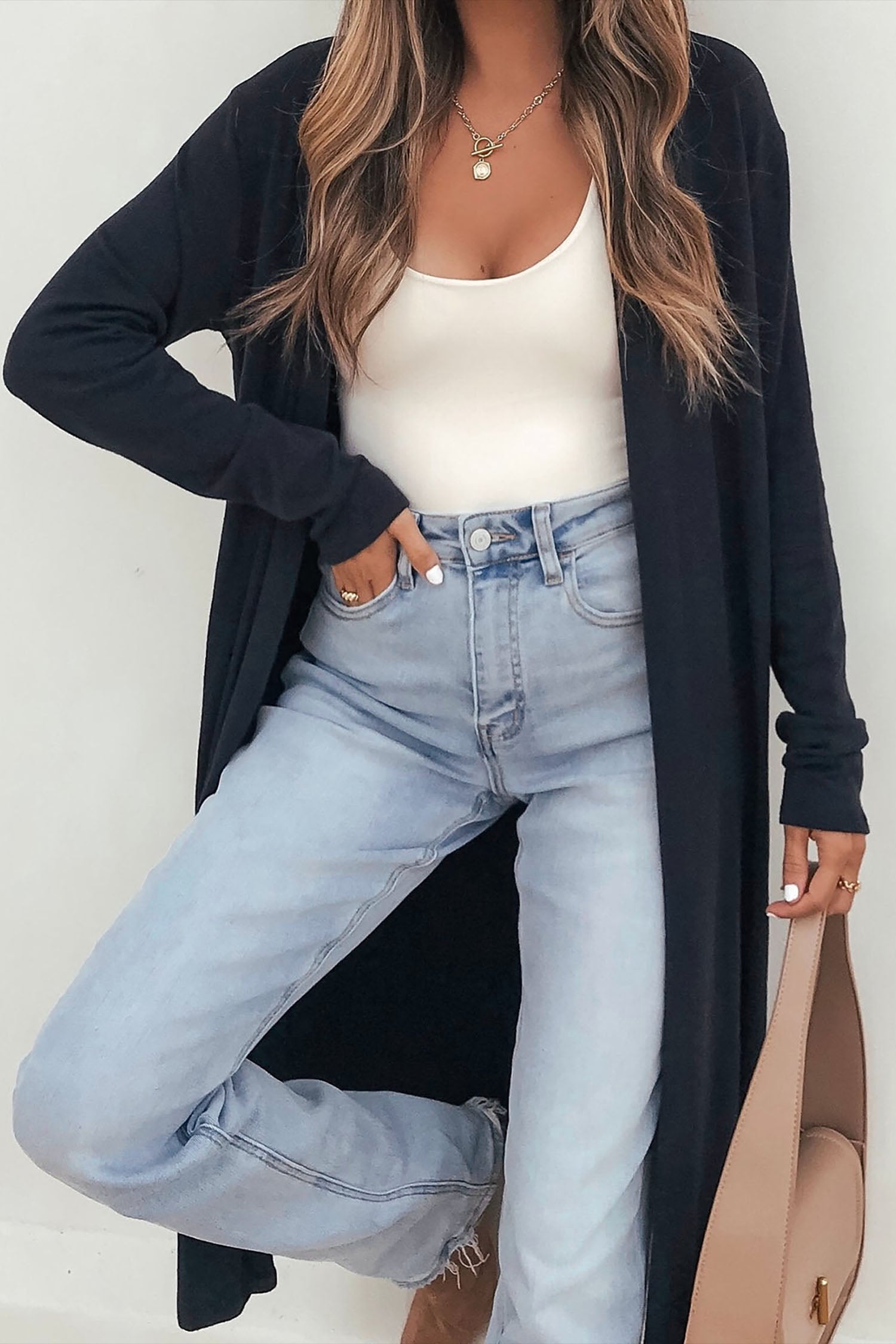 Woman in long black cardigan, white ribbed tank, blue jeans, and beige handbag.