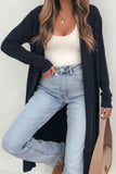 Woman in long black cardigan, white ribbed tank, blue jeans, and beige handbag.