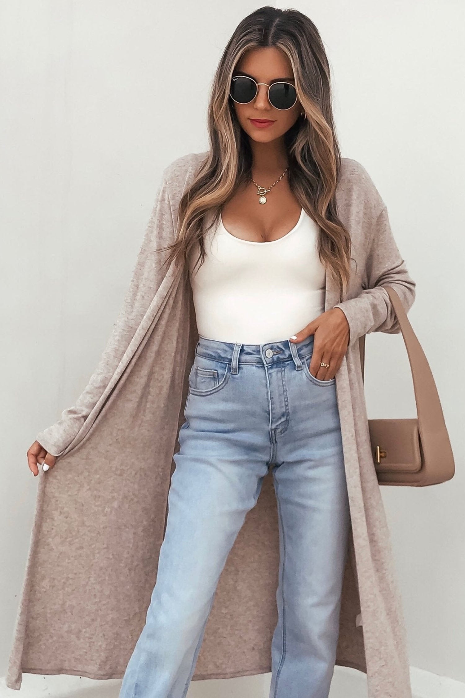 Woman in long beige cardigan, white tank, light blue jeans, sunglasses, and tan bag.