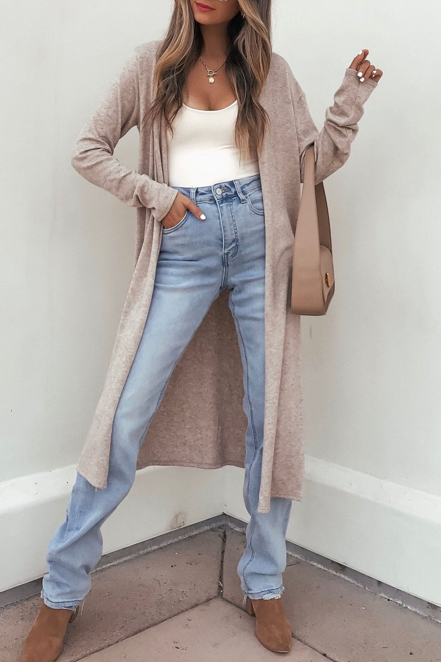 A woman modeling a long, open-front beige cardigan over a white tank top and light blue jeans