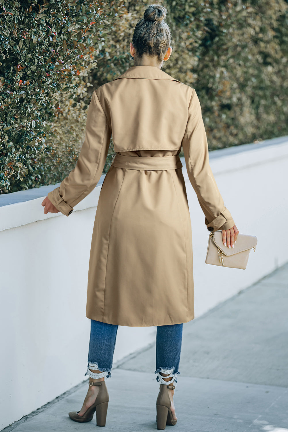 Person wearing a beige trench coat walking on a sidewalk.