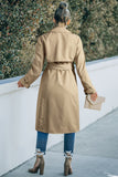 Person wearing a beige trench coat walking on a sidewalk.