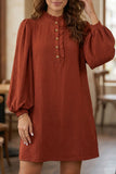 A woman wearing a rust-colored long-sleeve dress with a ruffled high neckline and gold buttons, shown in a softly lit indoor setting.