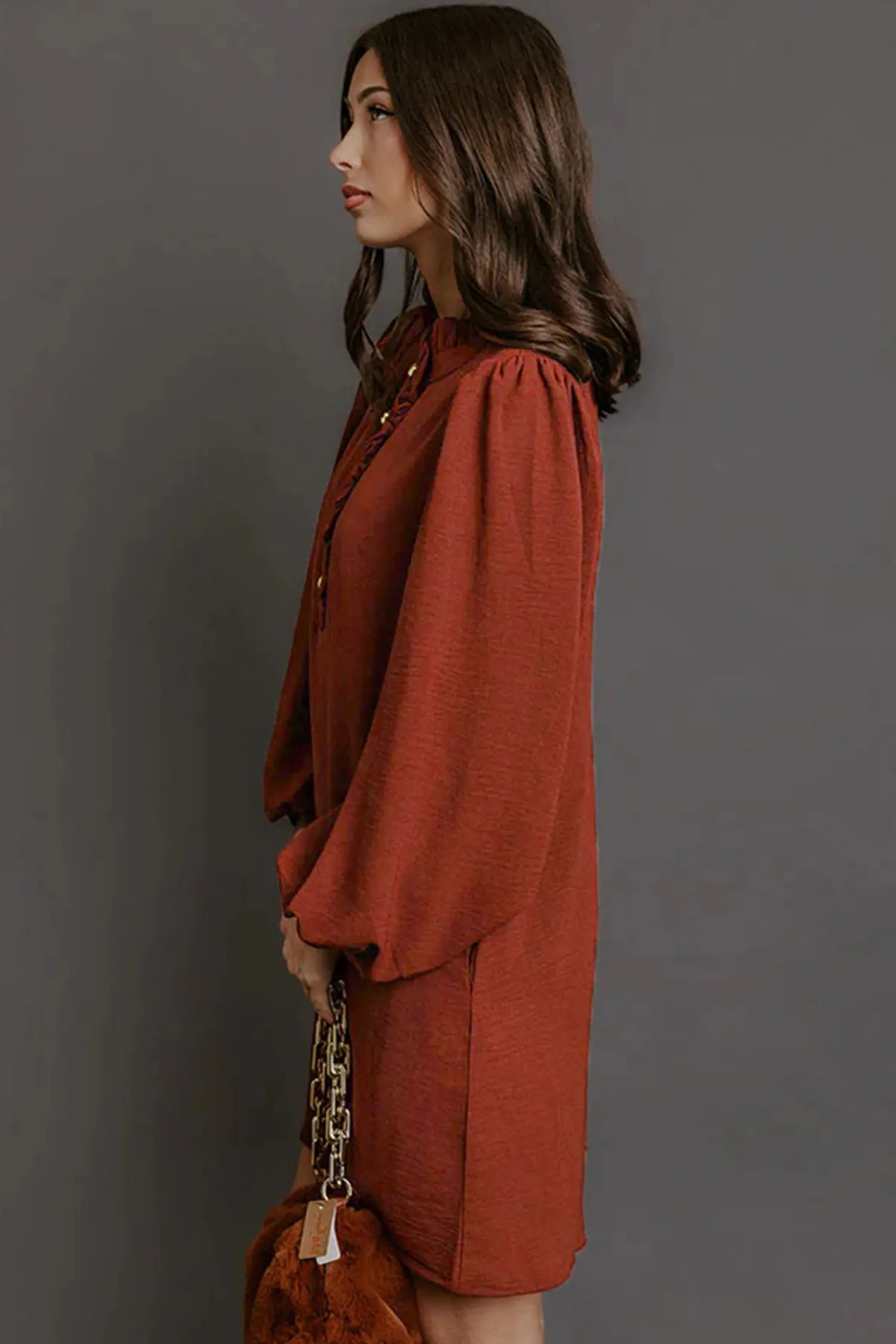 A woman in side profile wearing a rust-orange long-sleeve blouse with puffed shoulders, holding a brown handbag with a gold chain strap, set against a solid dark gray background.