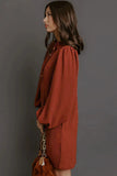 A woman in side profile wearing a rust-orange long-sleeve blouse with puffed shoulders, holding a brown handbag with a gold chain strap, set against a solid dark gray background.
