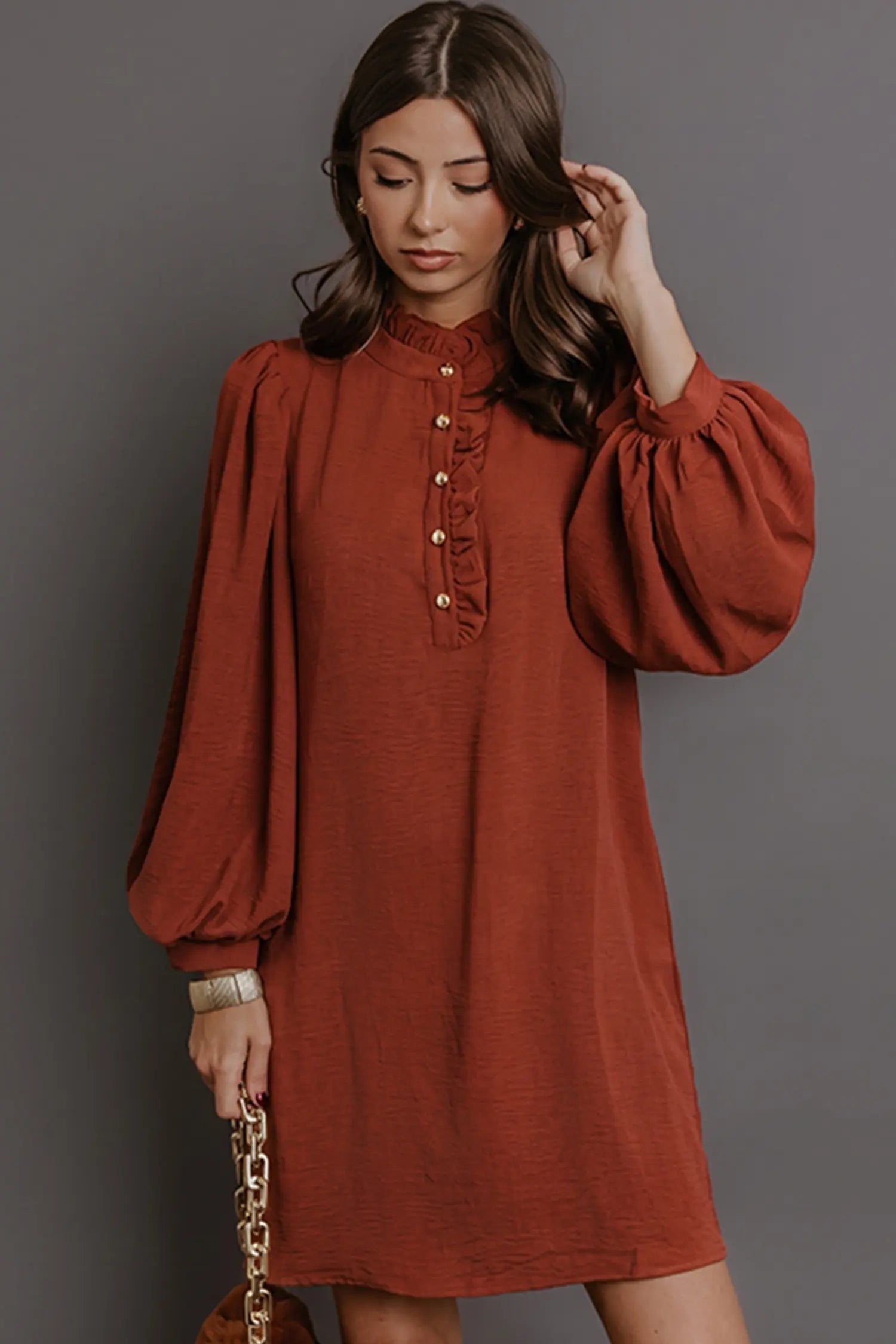 A woman wearing a rust-orange long-sleeve dress with a high ruffled collar and gold buttons, posing against a solid gray background.