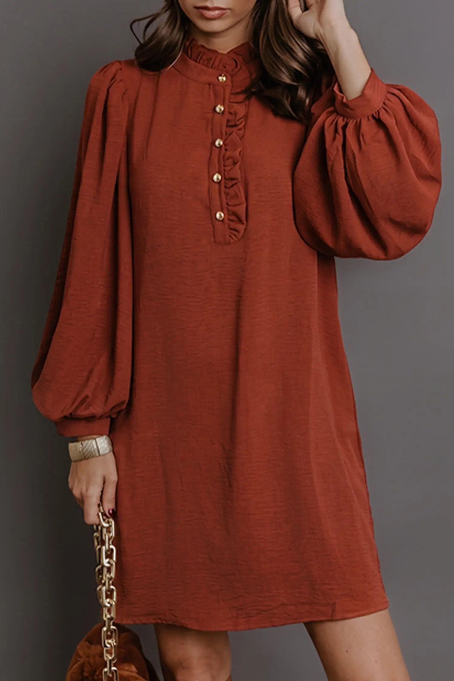 A woman wearing a rust-orange long-sleeve dress with a ruffled mock neck and decorative buttons down the front, paired with a gold chain handbag against a dark gray background.