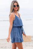 Woman in a denim dress standing on a beach with sunglasses and a straw bag.