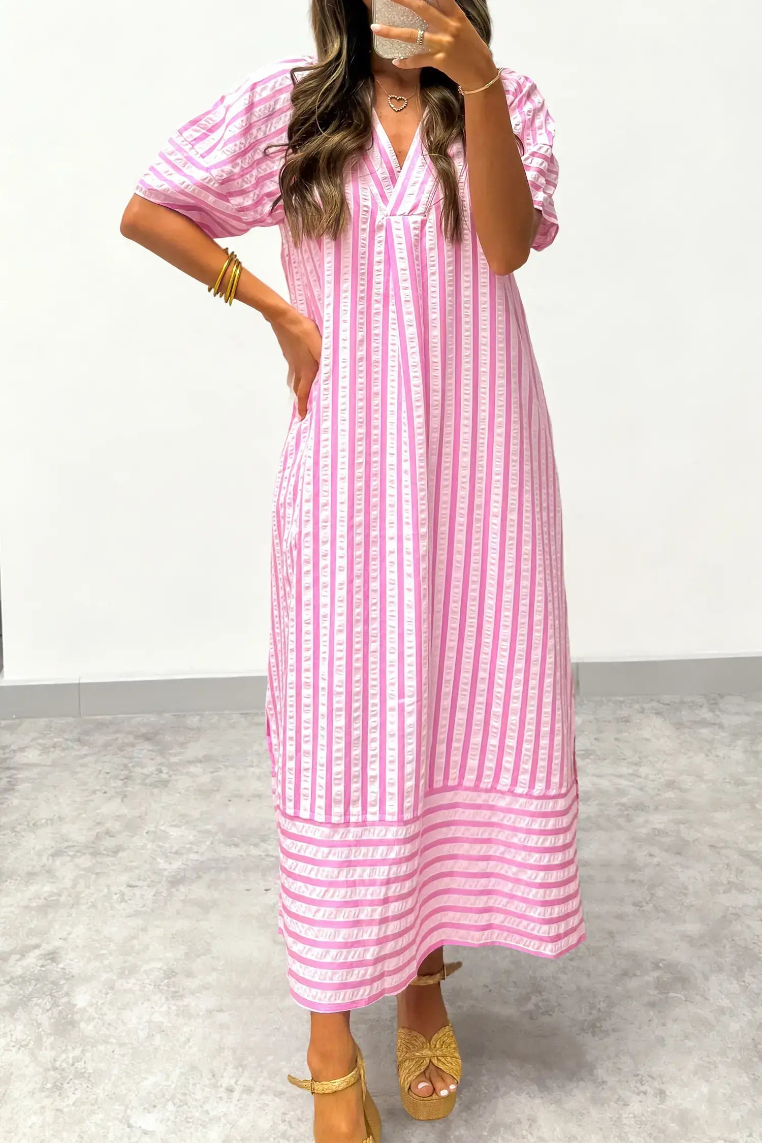 A woman wearing a pink and white vertically striped midi dress with puff sleeves and a V-neckline, paired with gold sandals and bracelets, taking a mirror selfie.