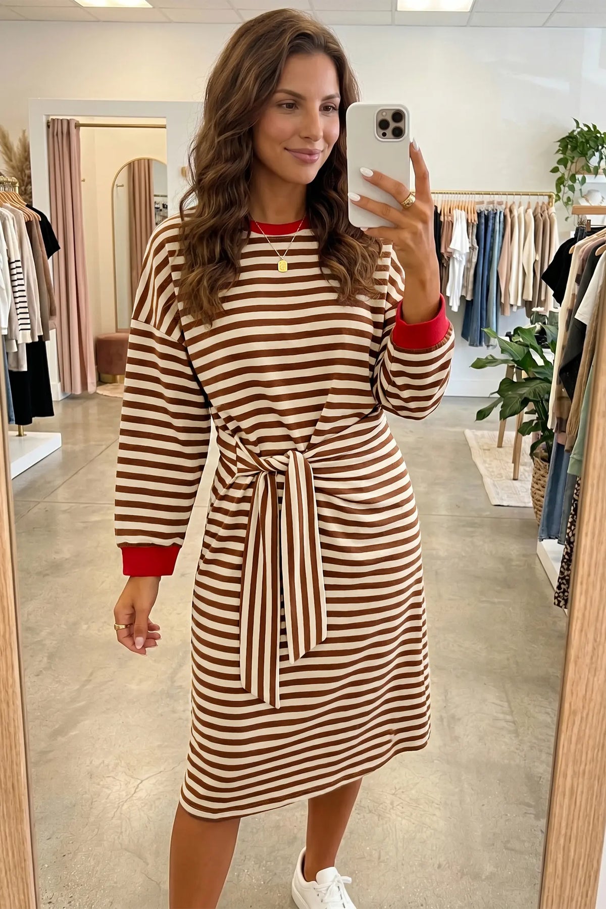 A woman takes a mirror selfie in a clothing boutique wearing a brown and cream striped long-sleeve dress with red cuffs and a red crew neck, paired with white sneakers.