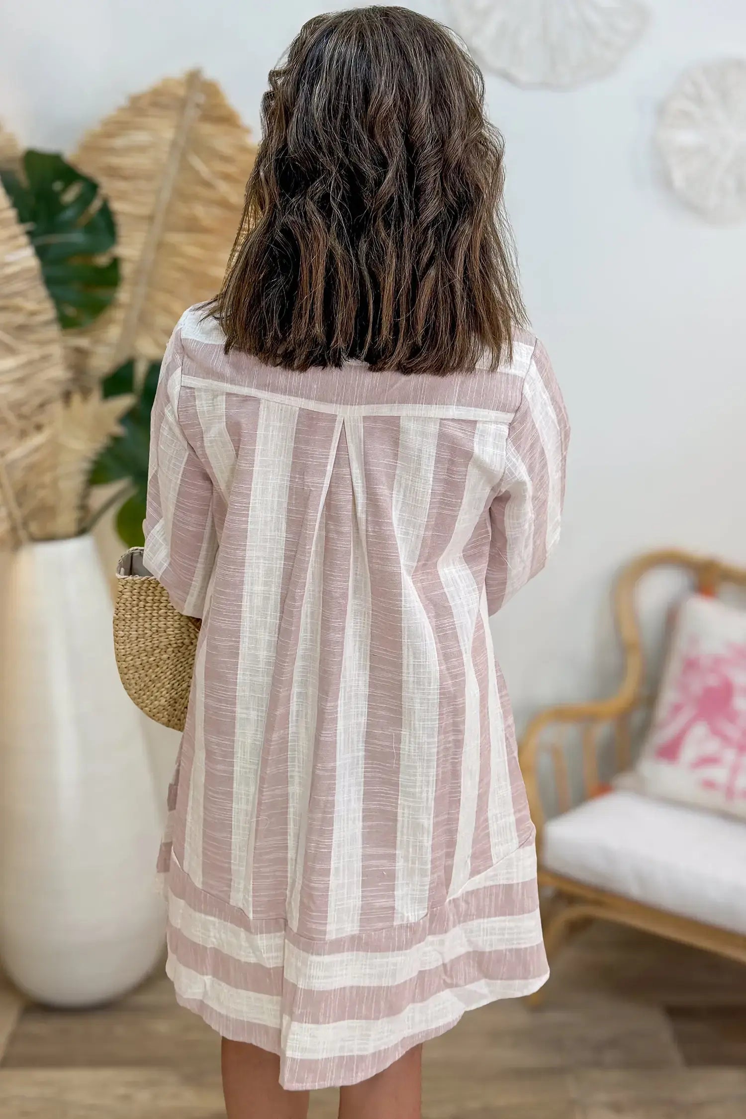 A child viewed from behind wearing a loose, short-sleeve striped tunic or dress in dusty pink and off-white, standing in a bright indoor room with woven decor and a chair in the background.