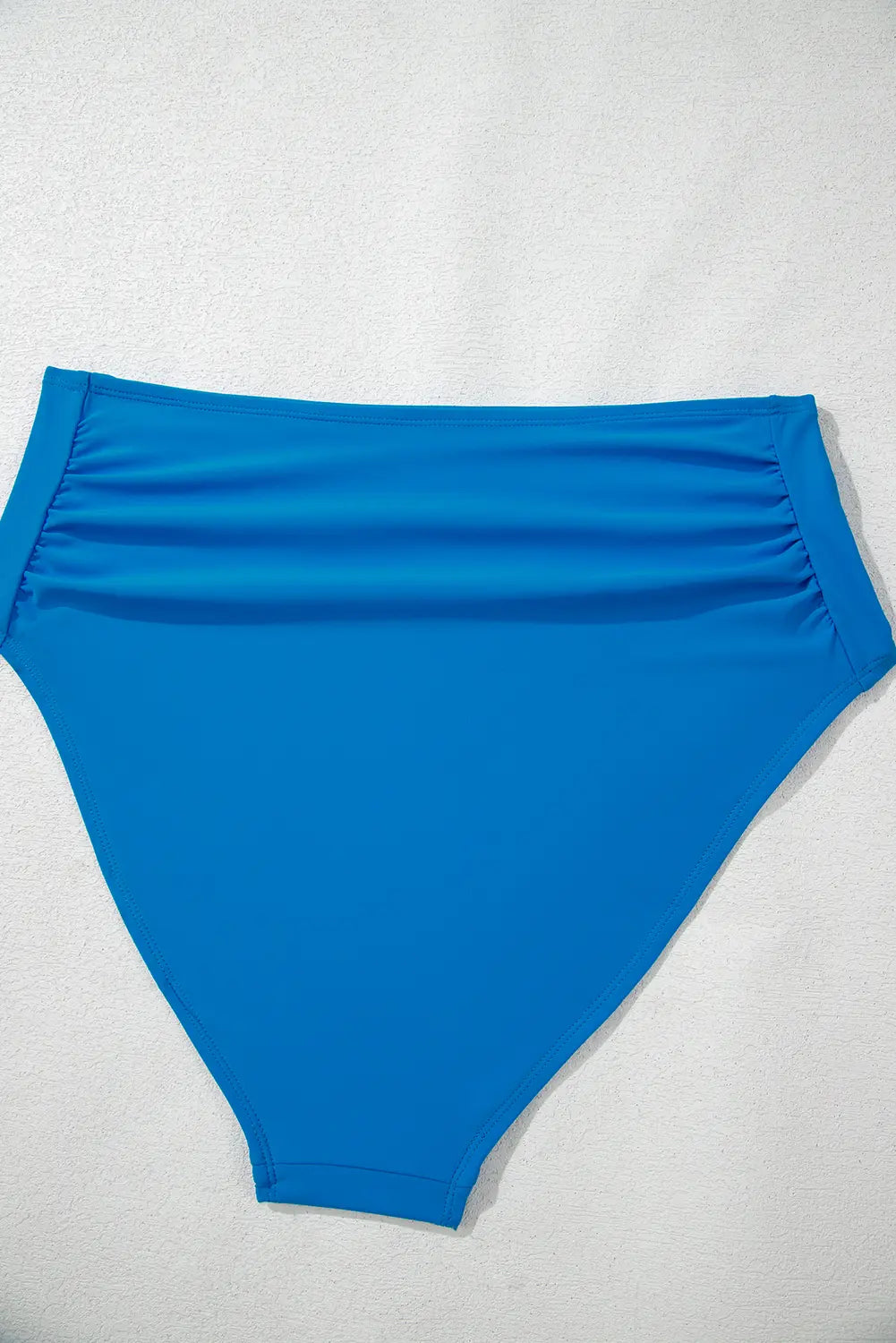 A bright blue high-waisted bikini bottom laid flat on a light gray textured background.