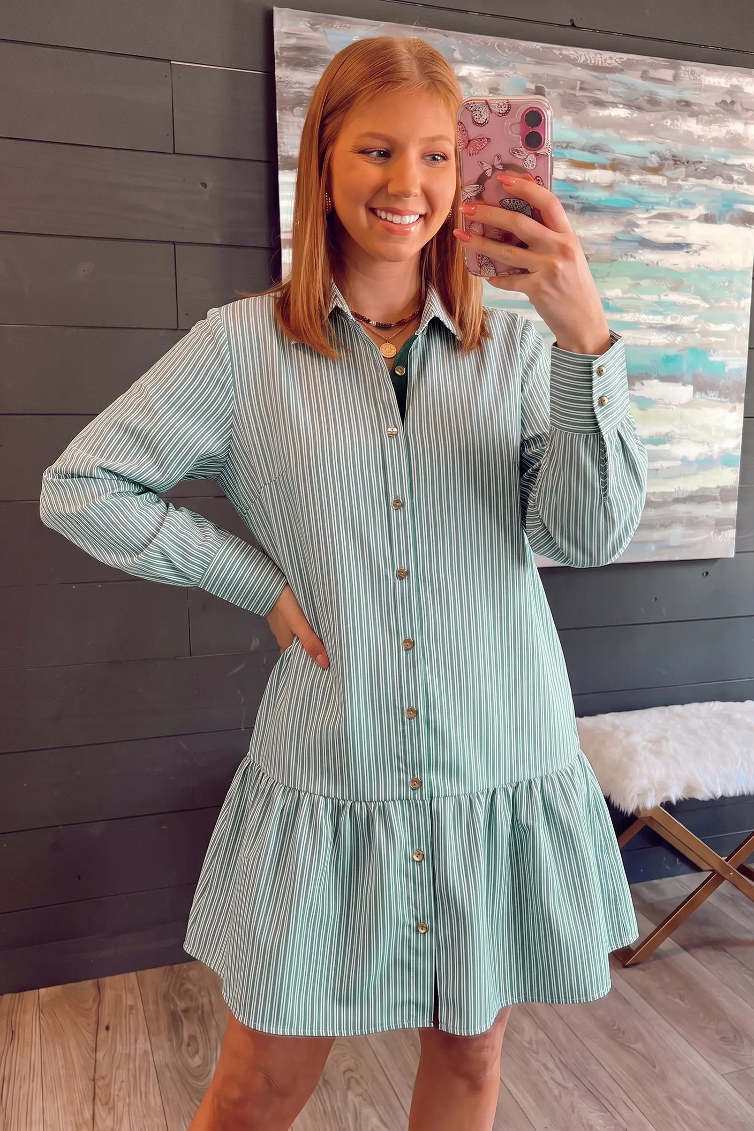 A woman takes a mirror selfie wearing a mint green and white vertically striped long-sleeve button-down shirt dress with a ruffled hem, set against a dark wood-paneled wall.