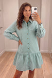 A woman takes a mirror selfie wearing a mint green and white vertically striped button-down shirt dress with a ruffled hem, standing in a fitting room with light wood floors.