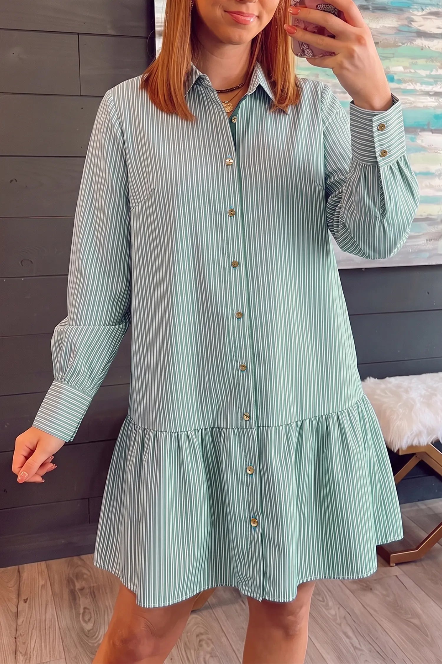 A woman wearing a mint green and white vertically striped long-sleeve shirt dress with gold buttons, taking a mirror selfie in an indoor setting.