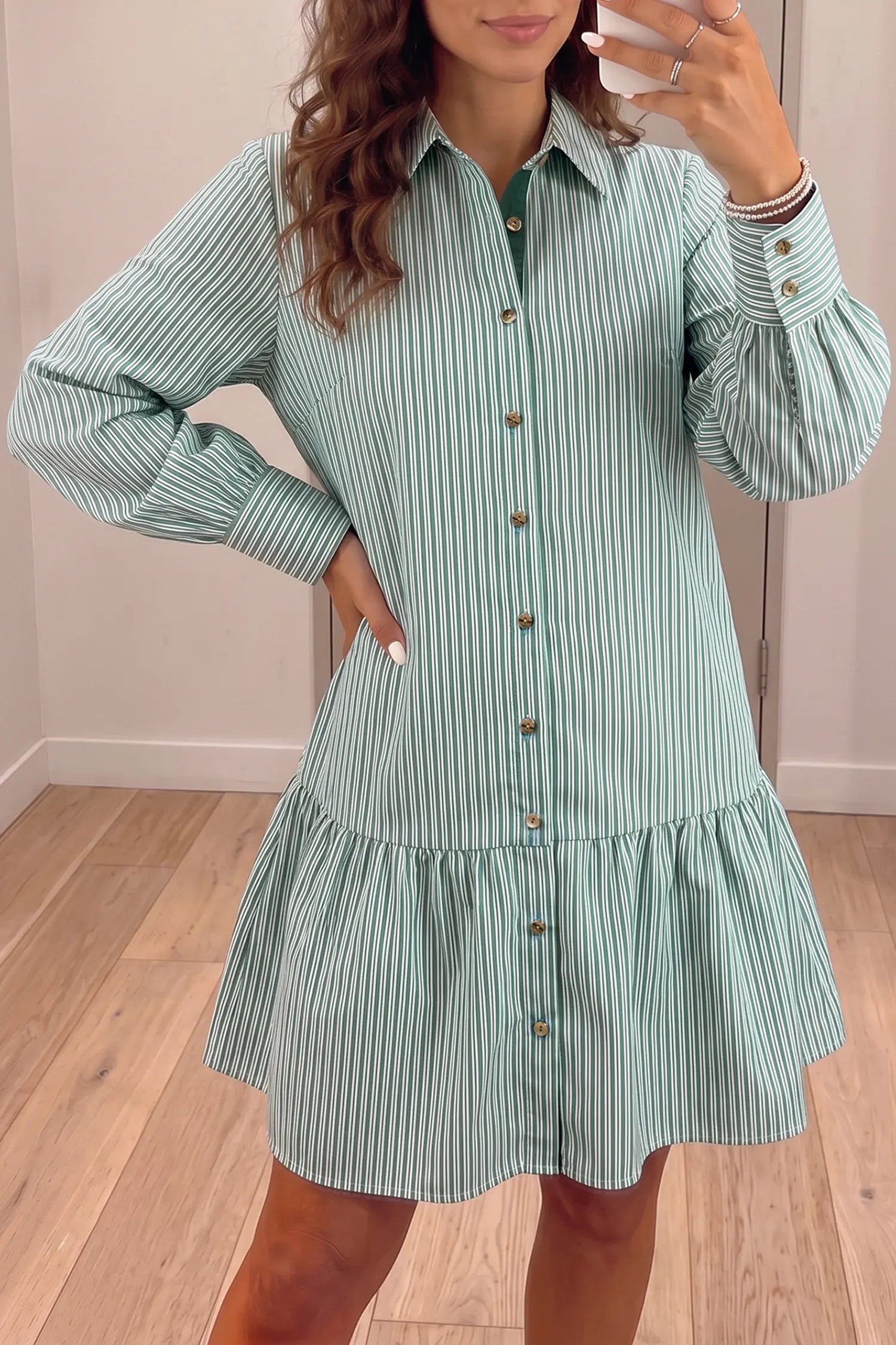 A woman wearing a mint green and white vertically striped button-down shirt dress, taking a mirror selfie in an indoor room with light wood floors.