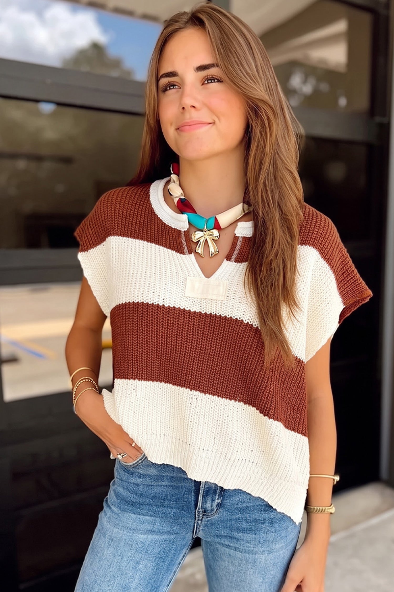 Woman in a brown and cream striped sweater vest with blue jeans and colorful jewelry.