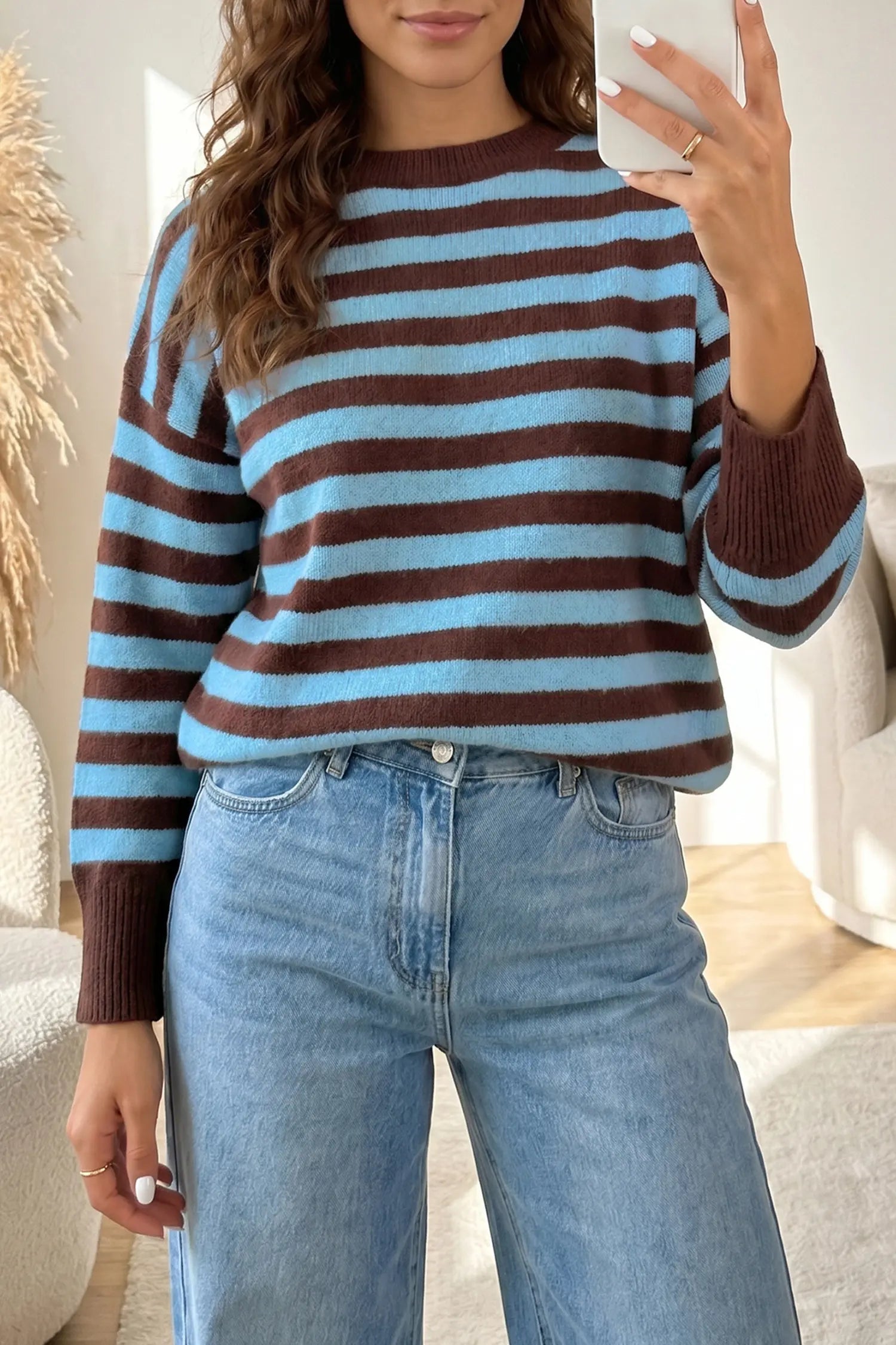 A woman takes a mirror selfie wearing a brown and light blue striped knit sweater with ribbed cuffs and hem, paired with light-wash blue jeans in a bright indoor setting.