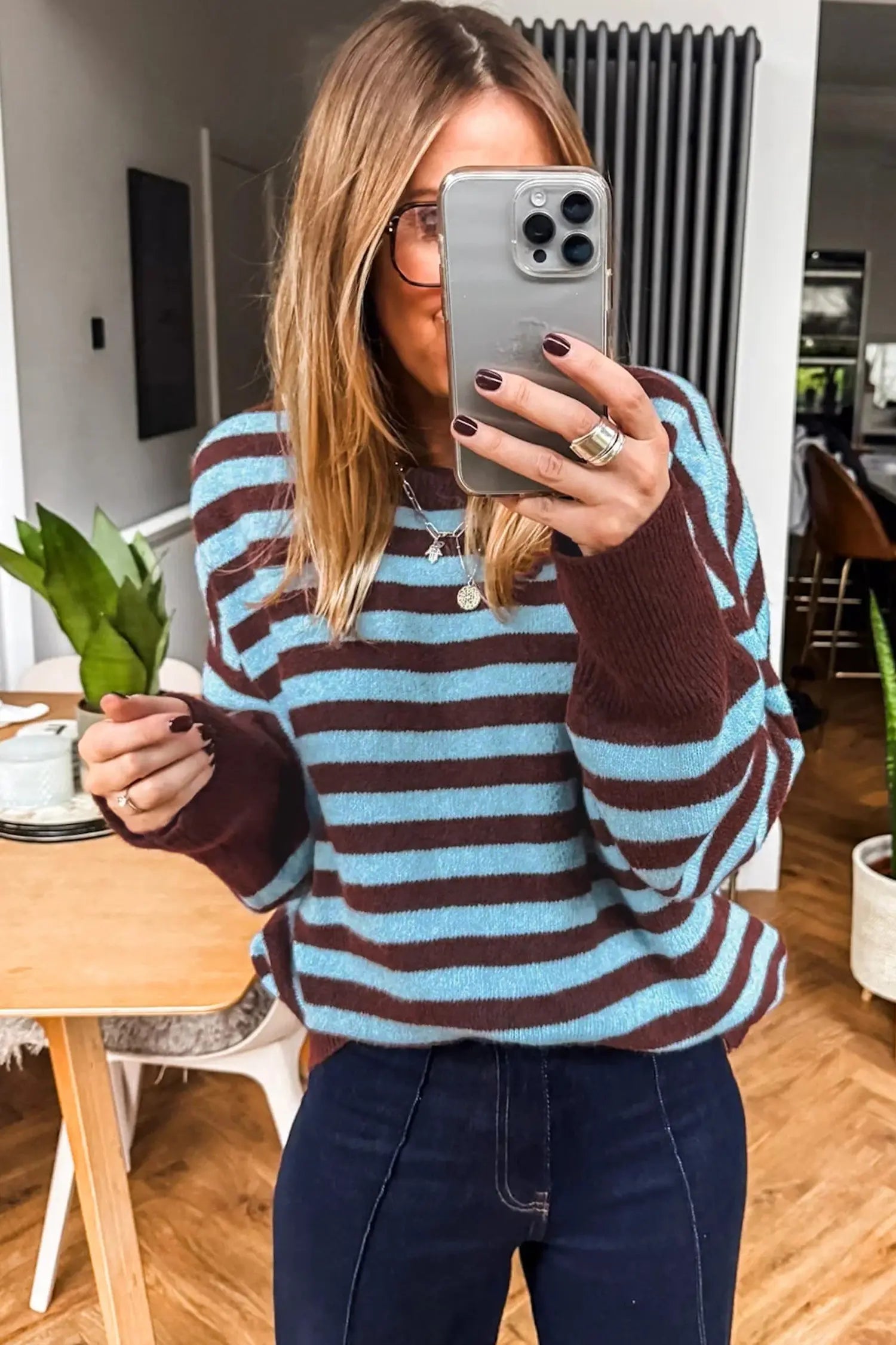 A woman takes a mirror selfie wearing a knit sweater with horizontal brown and light blue stripes, paired with dark blue jeans in a home interior setting.