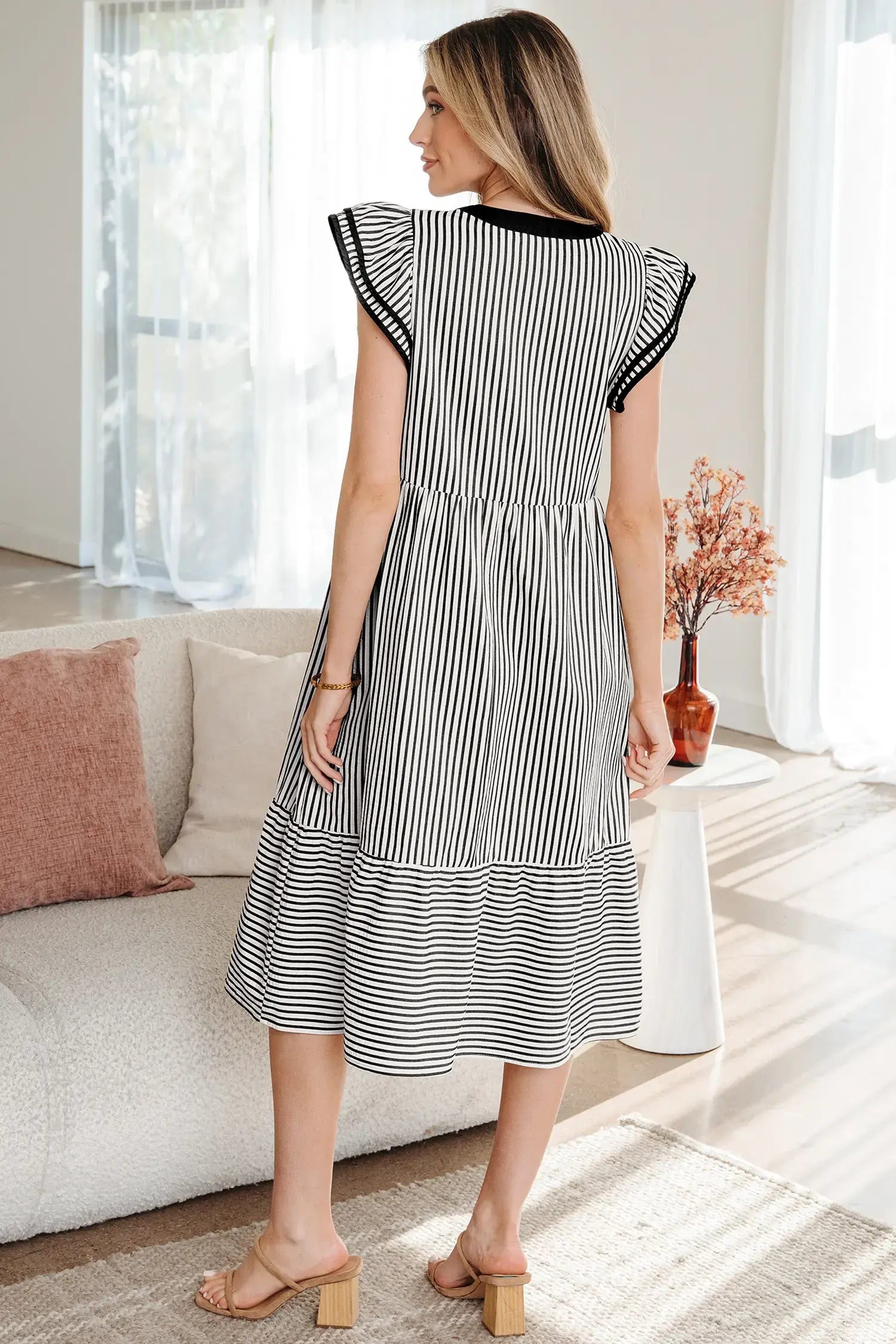 A woman wearing a black and white vertically striped midi dress with ruffled cap sleeves and black trim at the neckline, standing in a bright living room.