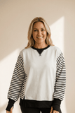 Woman wearing a white sweatshirt with black striped sleeves against a beige background