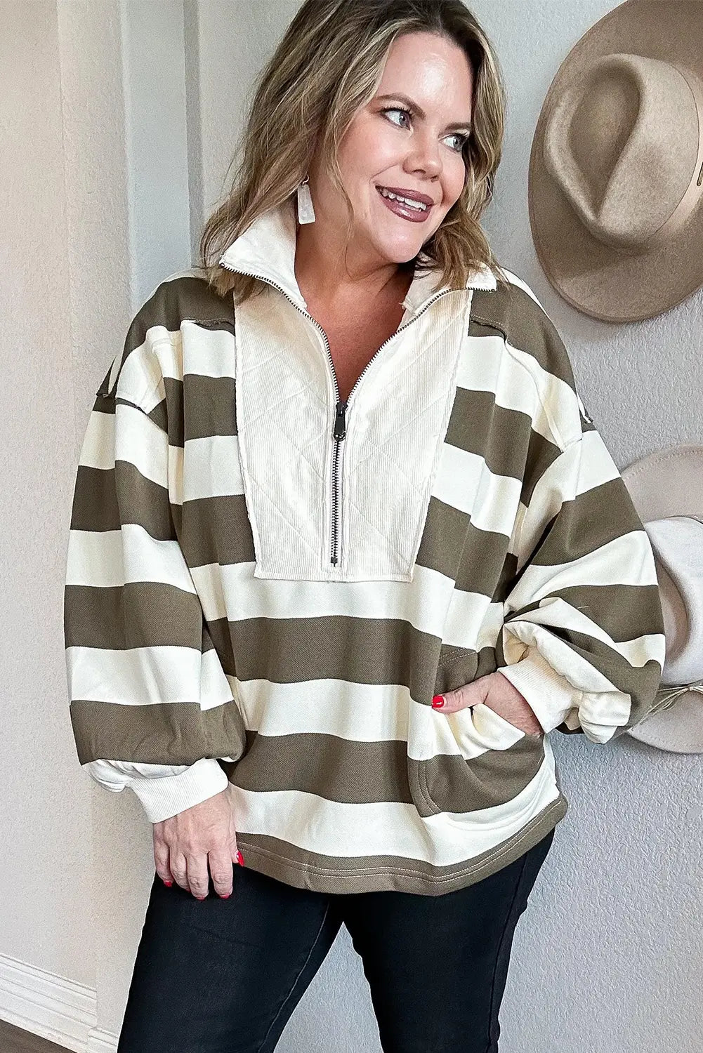 A woman wearing a cream and olive green horizontally striped quarter-zip pullover sweatshirt, paired with black pants, standing against a light-colored wall with hats hanging on the right.