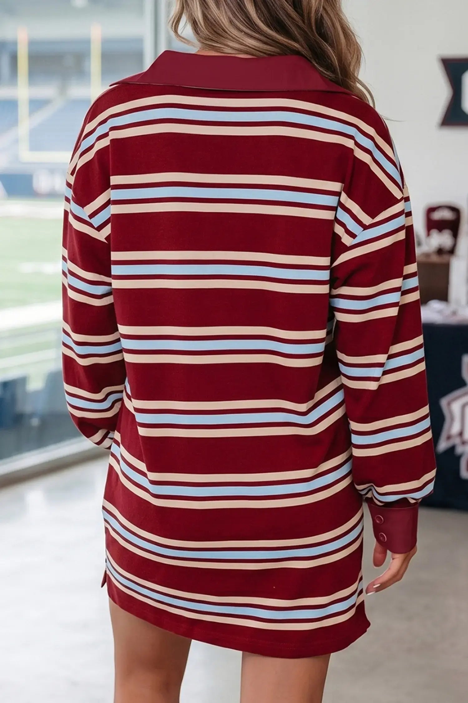 A woman viewed from behind wearing a long-sleeve collared polo shirt dress with horizontal stripes in burgundy, cream, and light blue. The setting appears to be an indoor sports facility or stadium.