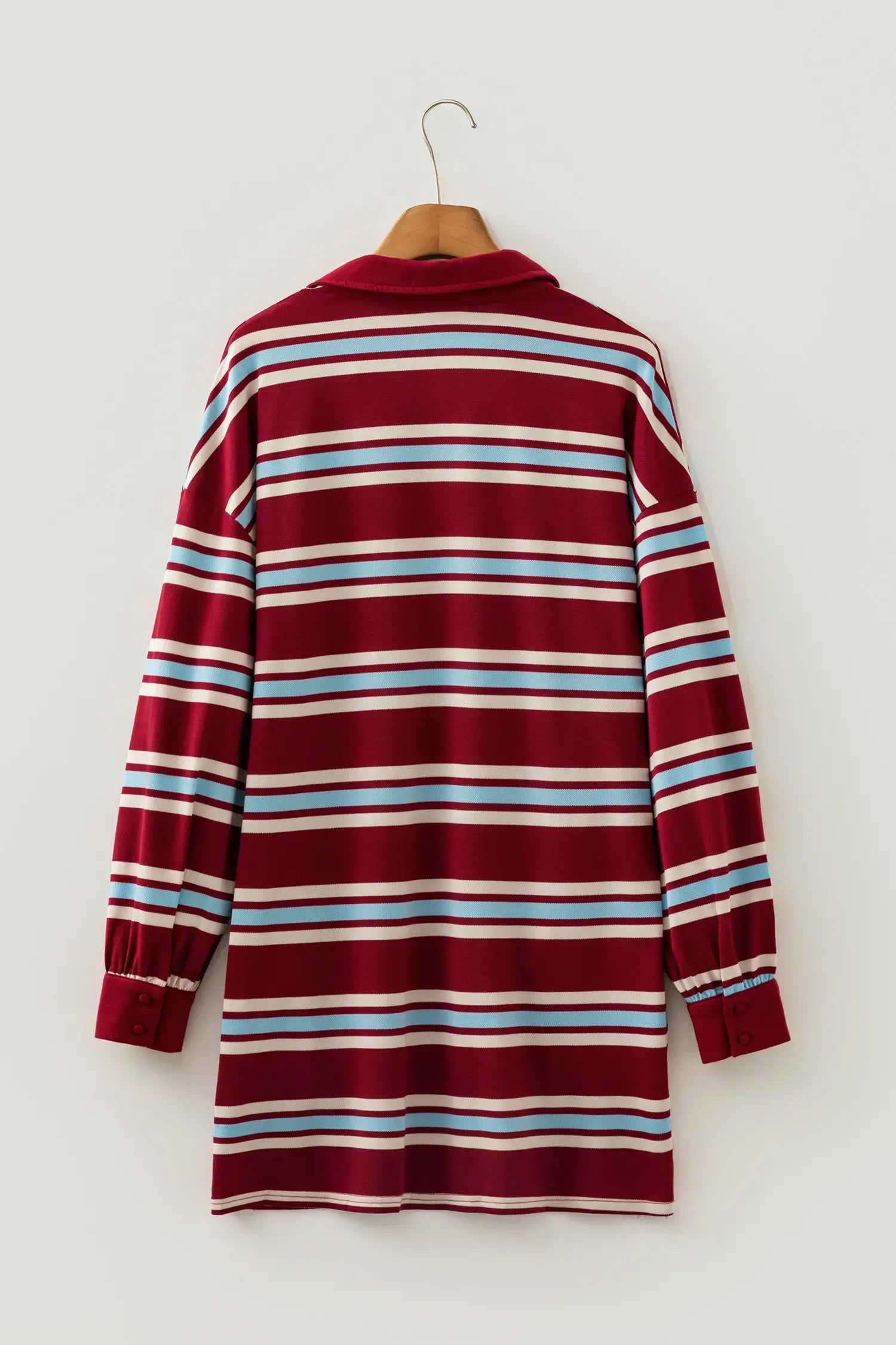 A long-sleeve collared shirt dress with horizontal stripes in burgundy, white, light blue, and thin red stripes, hanging on a wooden hanger against a light gray wall.