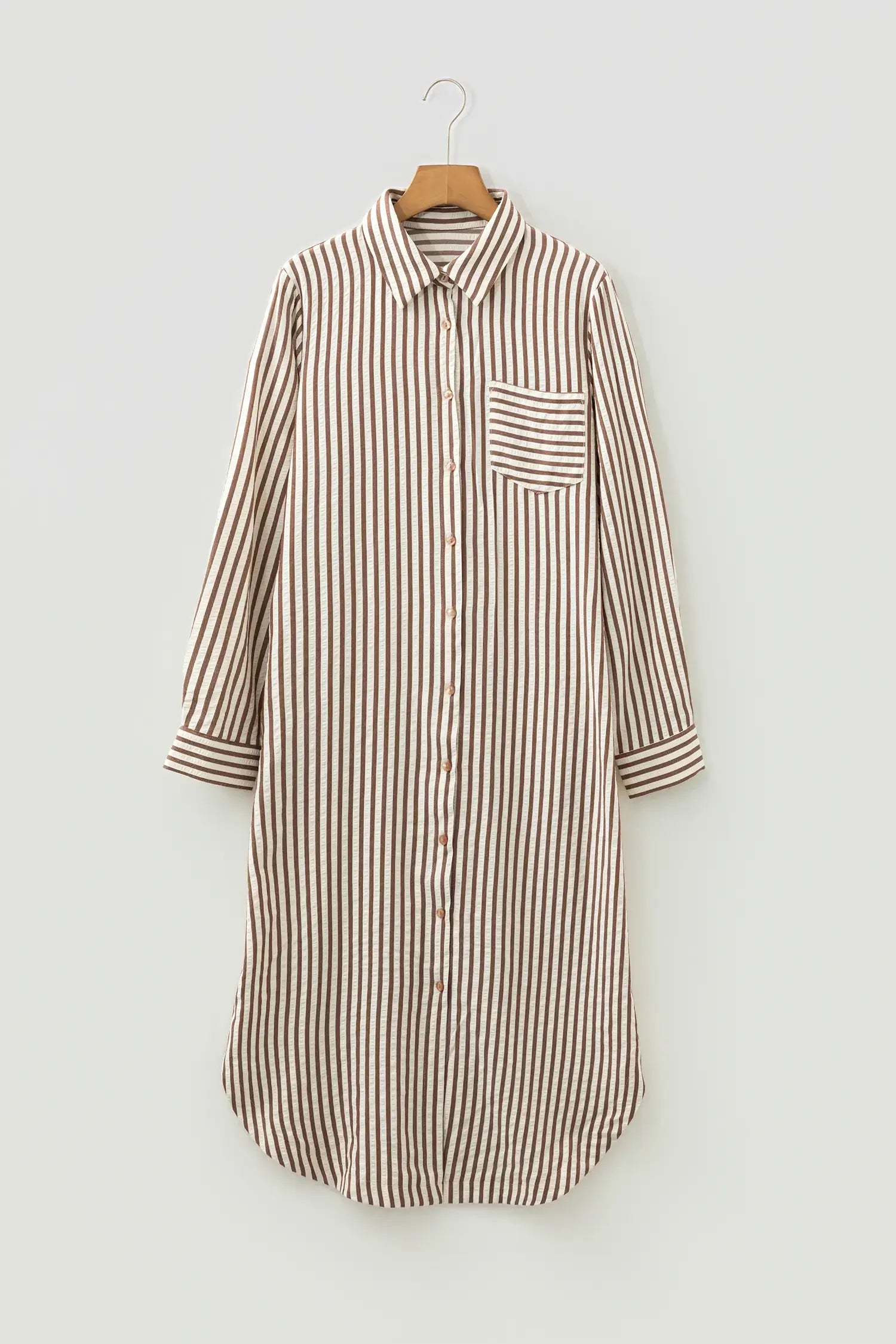 A long-sleeve button-front shirt dress with vertical brown and beige stripes, featuring a single chest pocket, shown hanging on a wooden hanger against a light gray background.