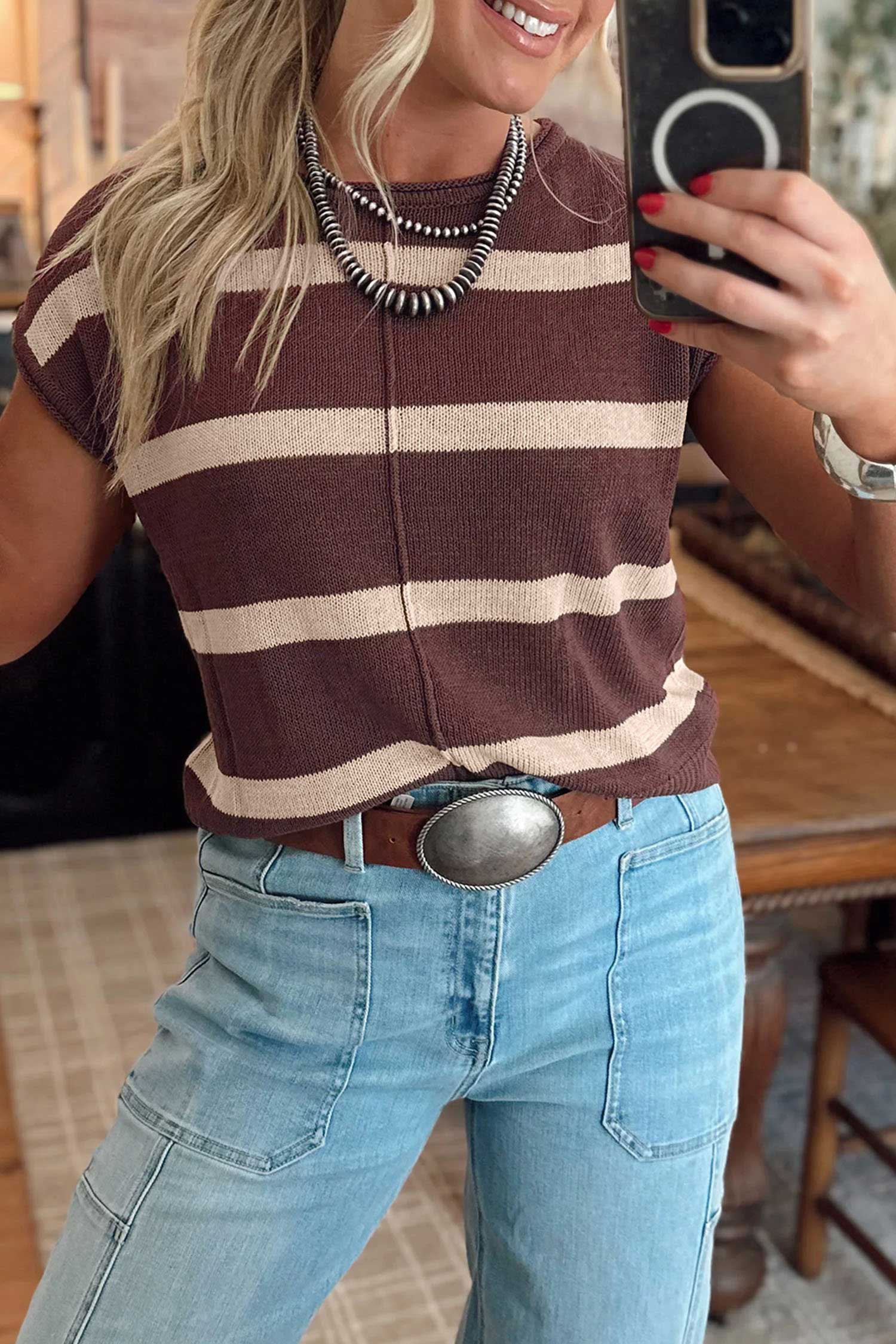 A woman takes a mirror selfie wearing a brown and beige horizontally striped short-sleeve knit top, light blue jeans, a brown belt with a large silver buckle, and layered silver necklaces.
