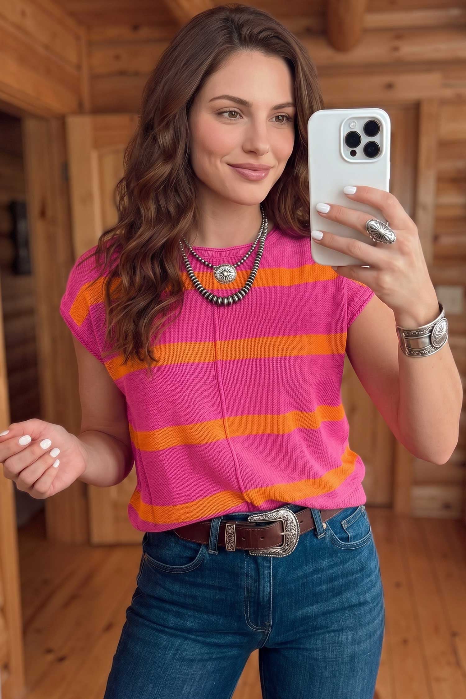 A woman takes a mirror selfie wearing a bright pink ribbed knit top with bold orange horizontal stripes, paired with blue jeans and a brown belt, in a rustic wood-paneled room.