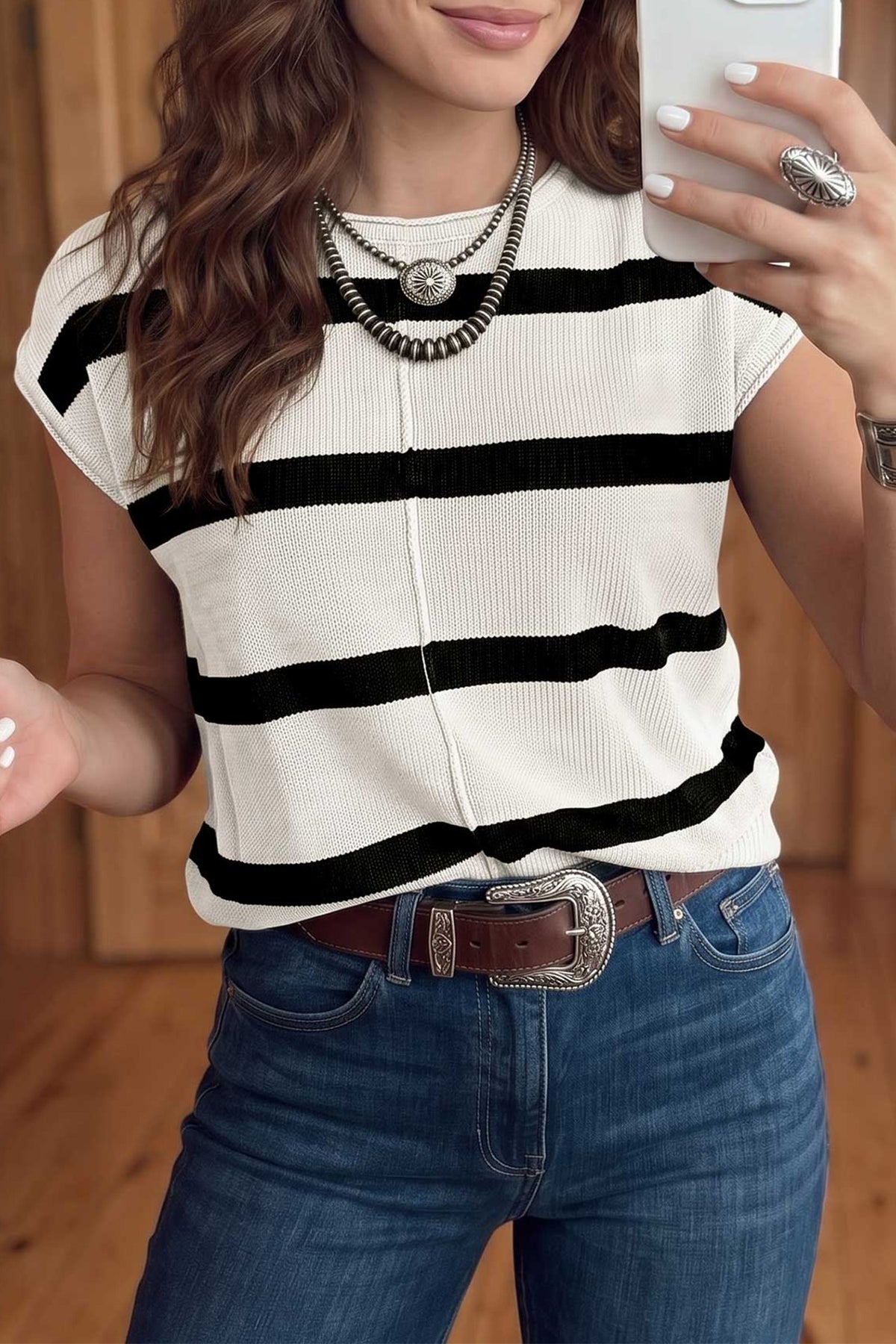A woman wearing a short-sleeve ribbed knit top with horizontal black and off-white stripes, paired with blue jeans and a brown belt, taking a mirror selfie.