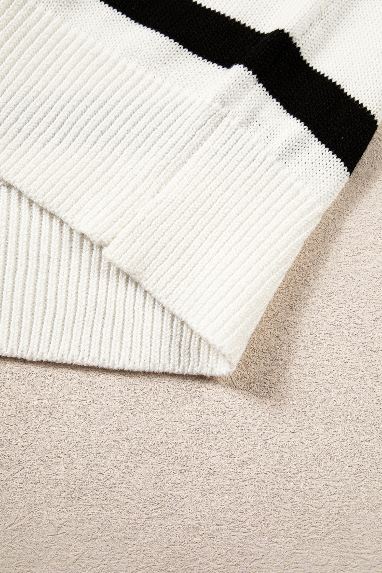 Close-up of a ribbed knit garment corner in off-white with a bold black stripe, laid on a textured light beige surface.