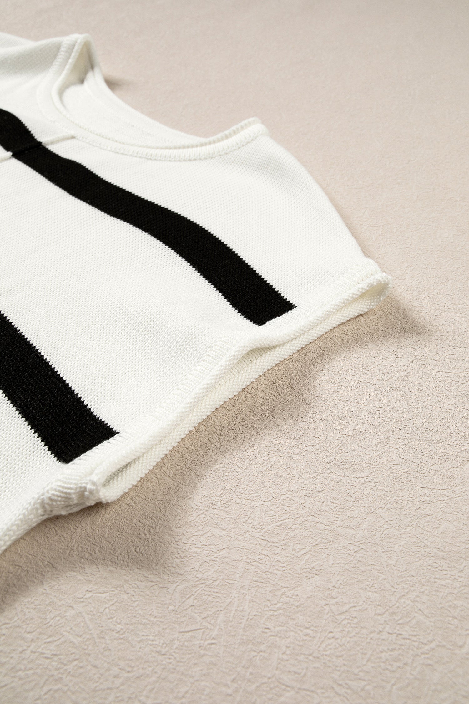 A close-up of a white knit garment with bold black diagonal stripes, laid flat on a textured beige surface.