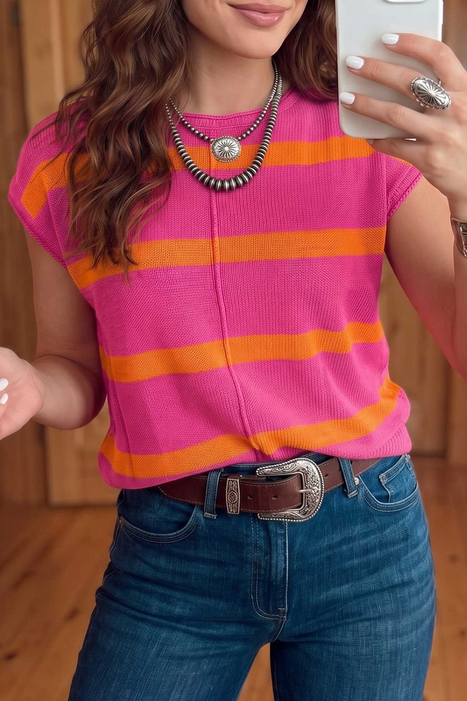 A woman wearing a bright pink short-sleeve knit top with bold orange horizontal stripes, paired with blue jeans and a brown belt, taking a mirror selfie.