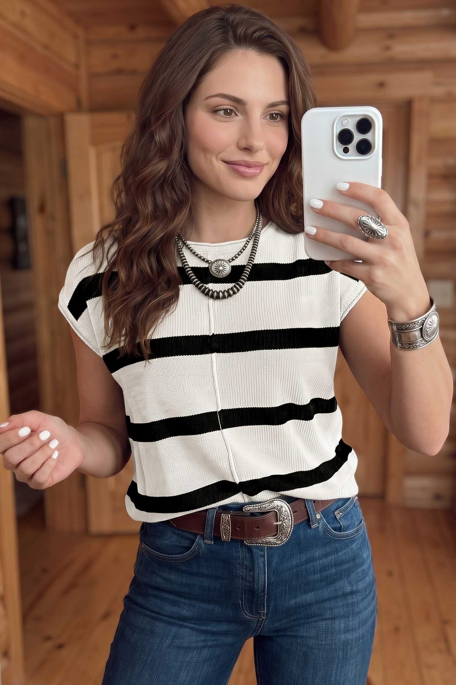 A woman takes a mirror selfie wearing a black and white horizontally striped ribbed knit top, blue jeans, and layered silver necklaces in a rustic wood-paneled room.