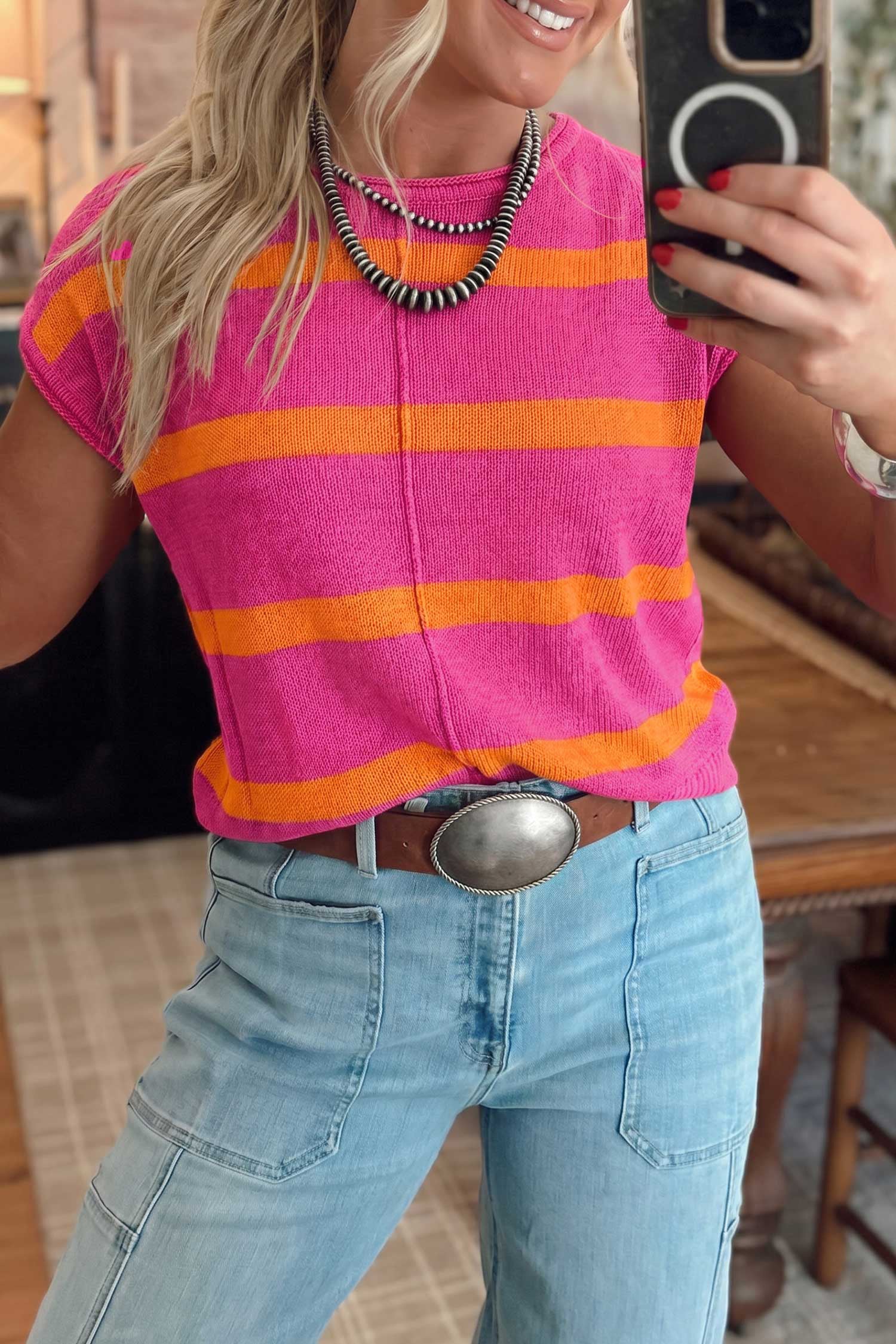 A woman takes a mirror selfie wearing a bright pink short-sleeve knit top with bold orange horizontal stripes, paired with light blue jeans and a brown belt with a large silver buckle.