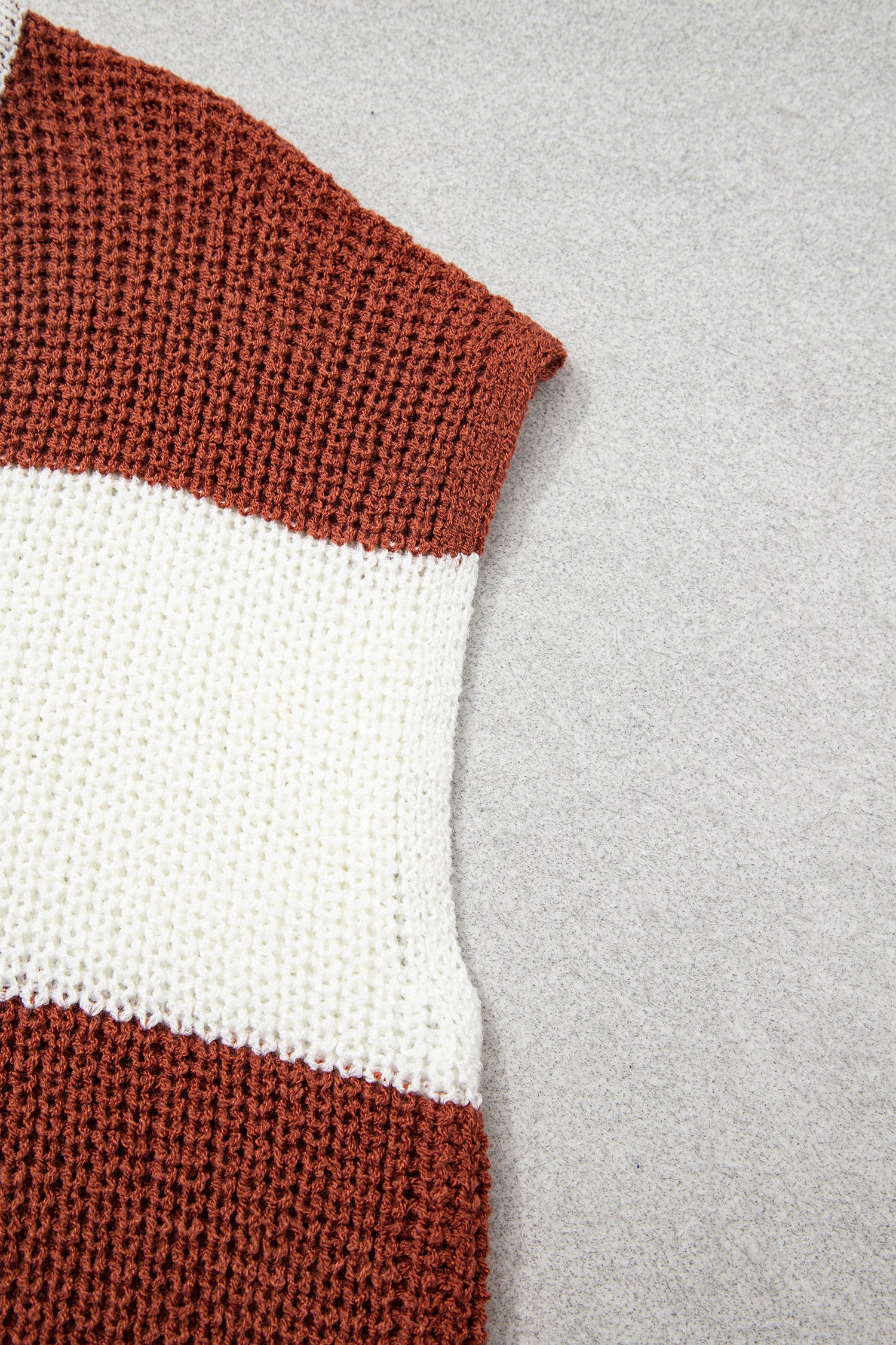 A close-up of a chunky knit sweater showing horizontal stripes in rust brown and off-white, laid flat on a light gray textured background.