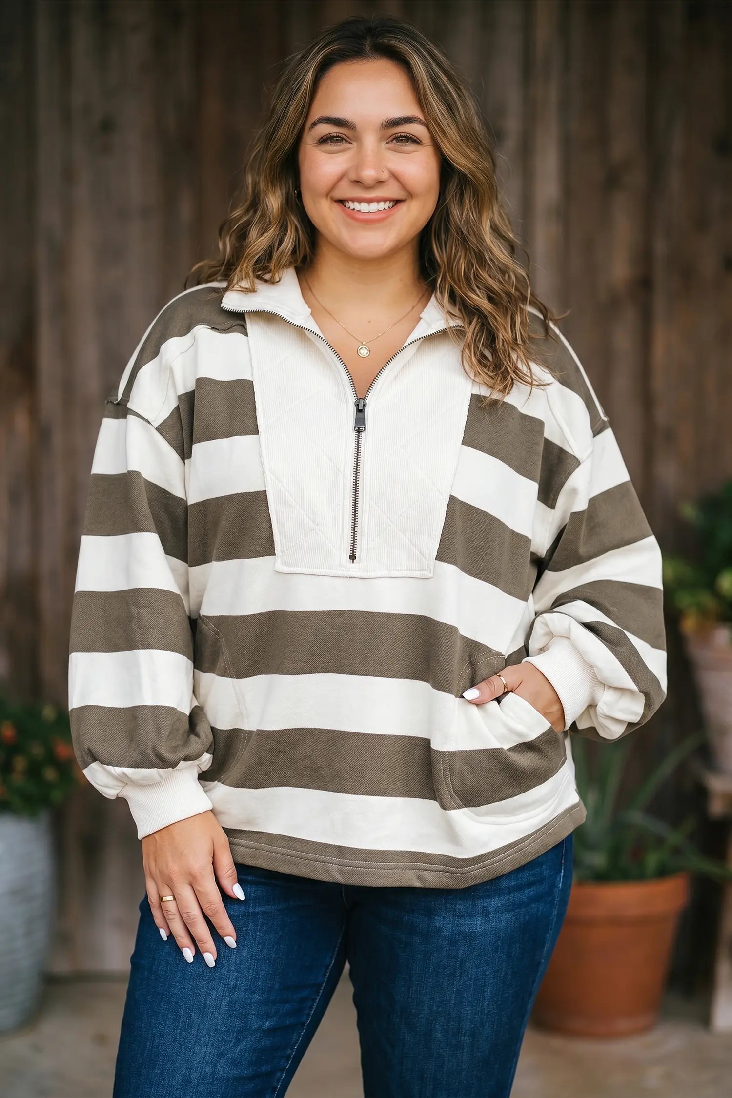 A woman wearing a brown and off-white horizontally striped quarter-zip pullover sweatshirt with pockets, paired with blue jeans, standing in front of a rustic wooden fence.
